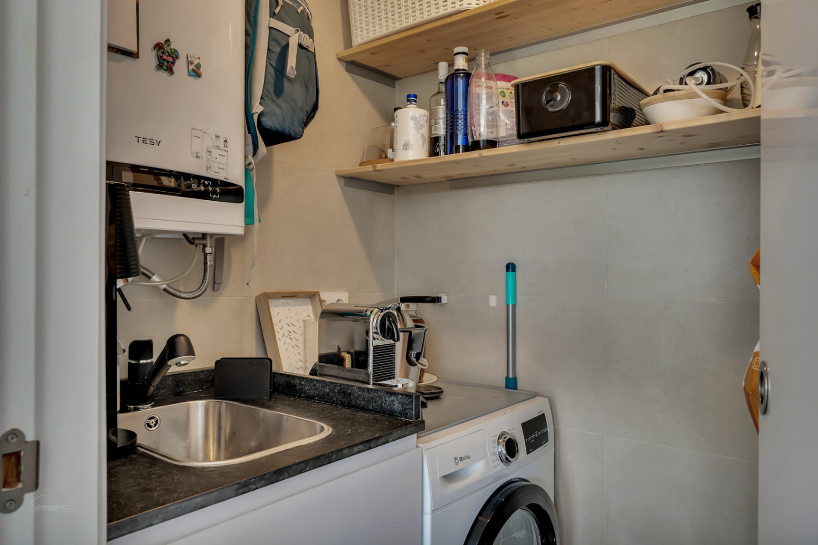 Funktionaler Hauswirtschaftsraum mit Balay-Waschmaschine, Edelstahlspüle, dunkler Arbeitsplatte und Regalen aus Naturholz an hellgrau gefliesten Wänden.