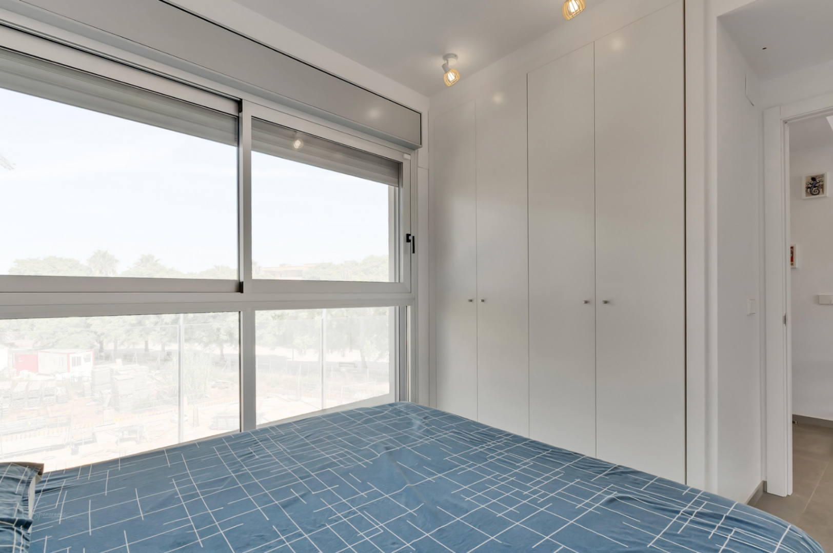 Helles Schlafzimmer mit deckenhohen weißen Einbauschränken, großen Schiebefenstern mit Blick ins Grüne und moderner Spotbeleuchtung.