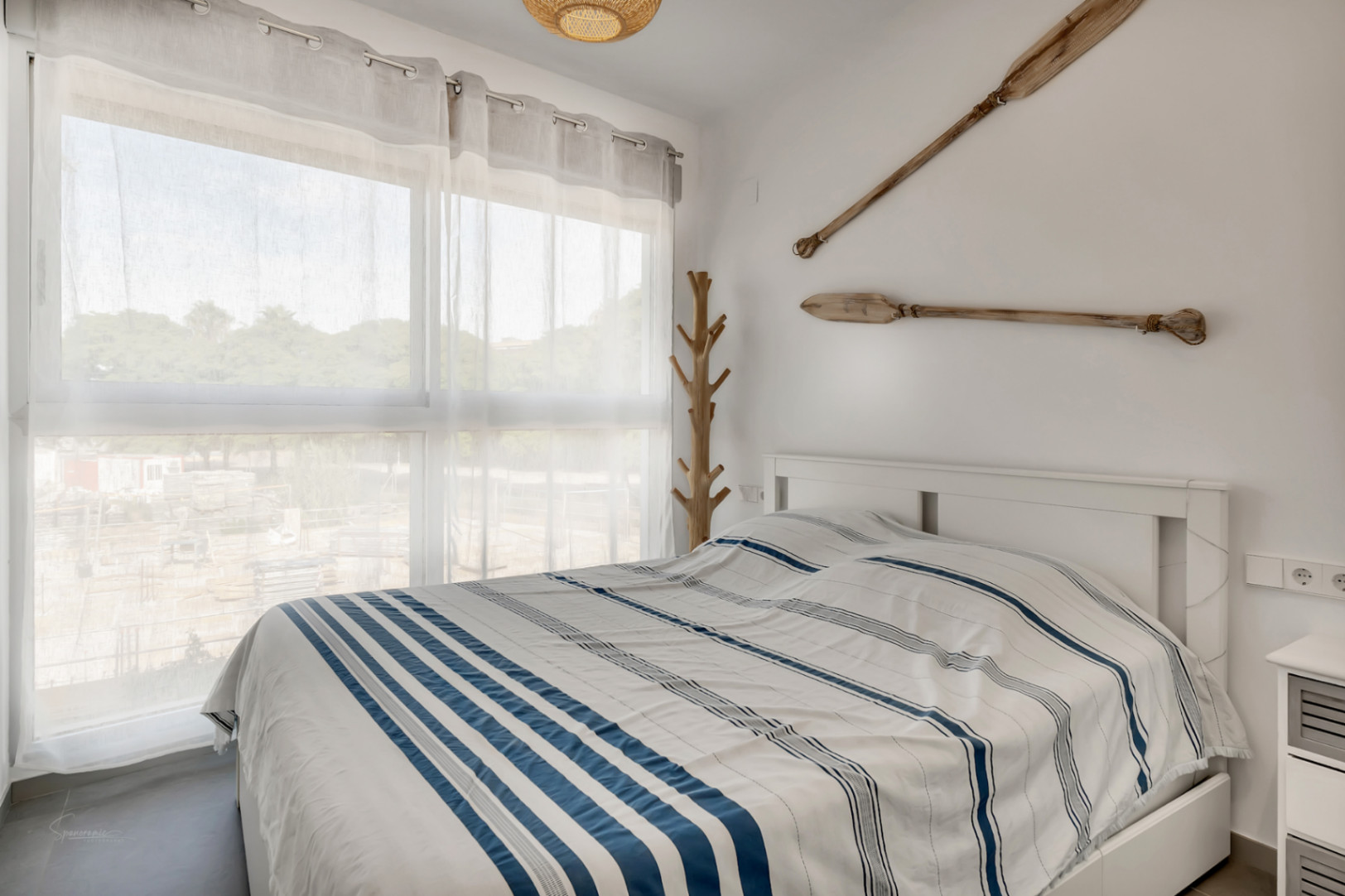 Helles Schlafzimmer mit Doppelbett, nautischer Dekoration aus Holzrudern und einem baumförmigen Garderobenständer neben dem großen Fenster.