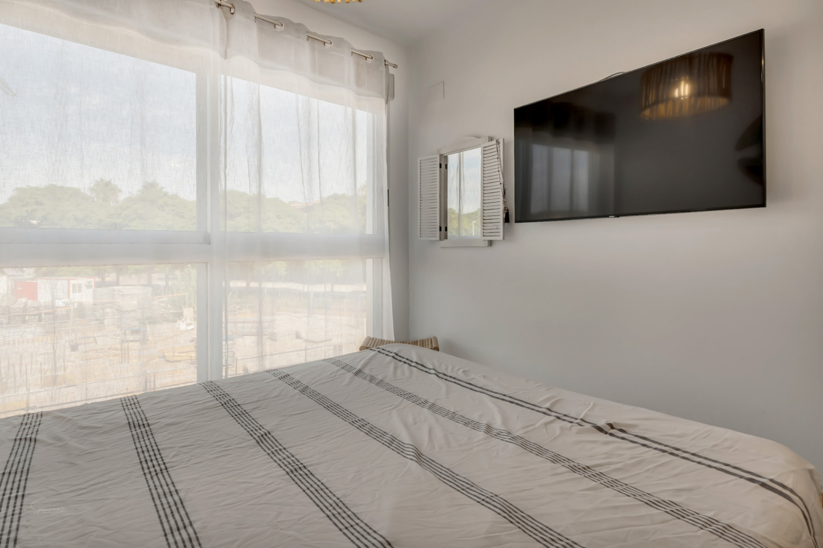 Helles Schlafzimmer mit großem Fenster, lichtdurchlässigen Vorhängen, Doppelbett mit gestreifter Bettwäsche und wandmontiertem Flachbildfernseher.