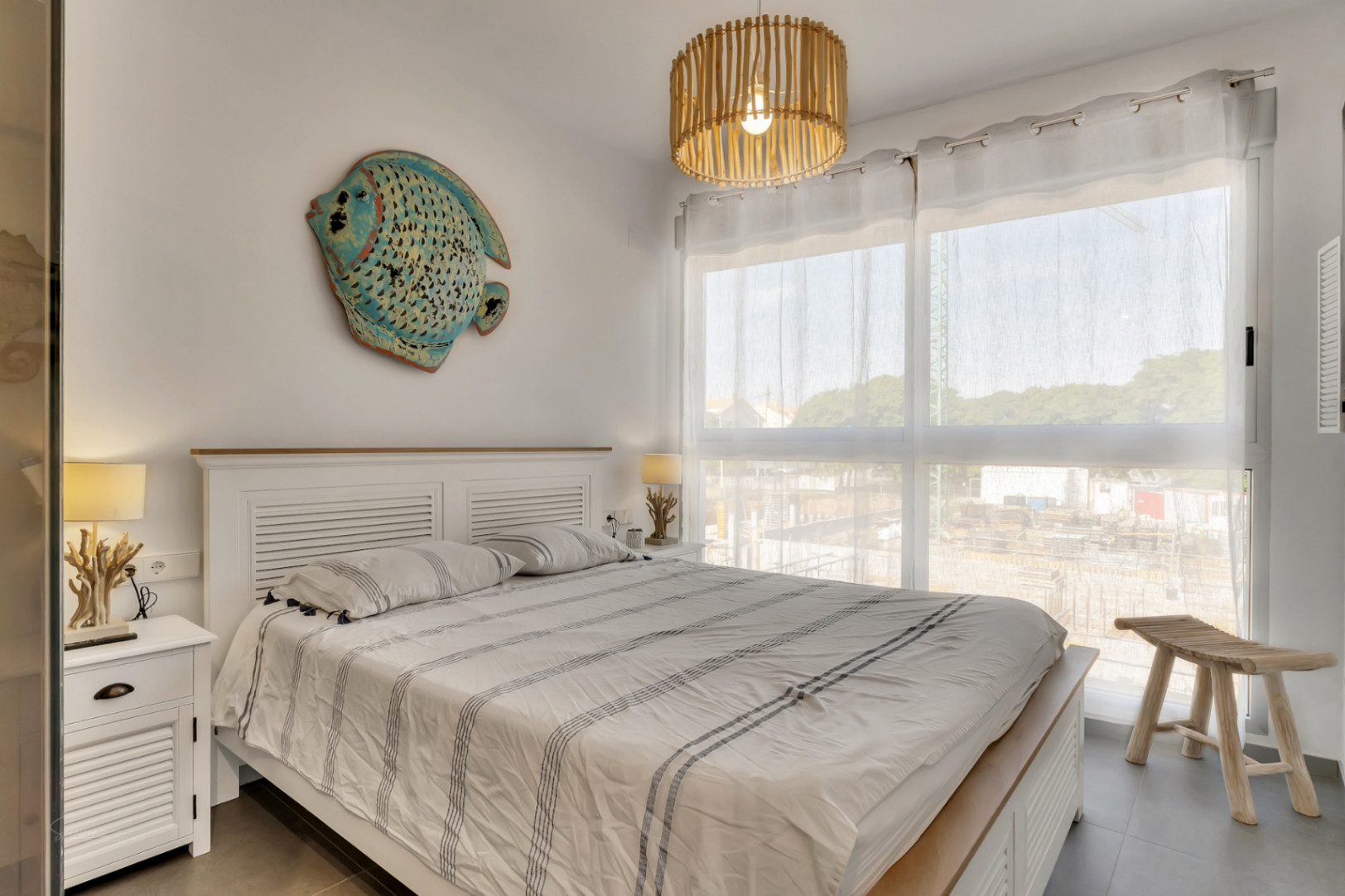 Helles Schlafzimmer mit weißem Holzbettrahmen, maritimer Dekoration, einer Bambus-Pendelleuchte und großem Fenster für natürliches Licht.
