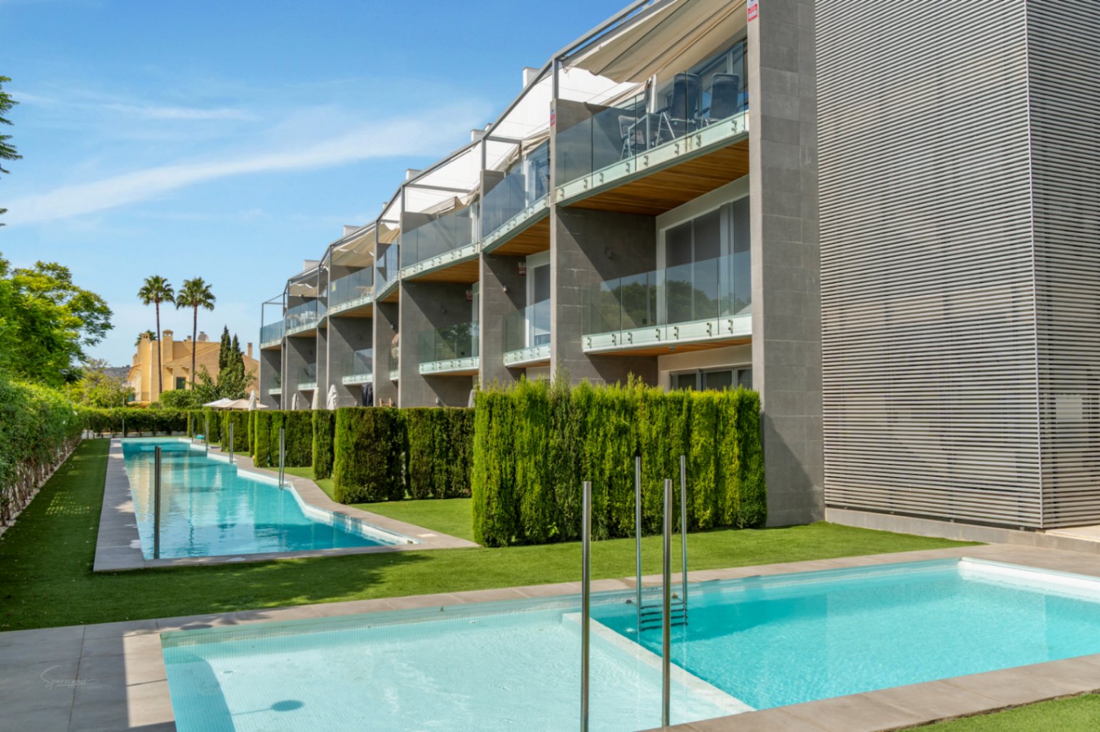 Moderne Fassade mit Glasbalkonen und Holzdetails, Blick auf den Gemeinschaftsbereich mit zwei Pools und gepflegten Heckenanlagen.