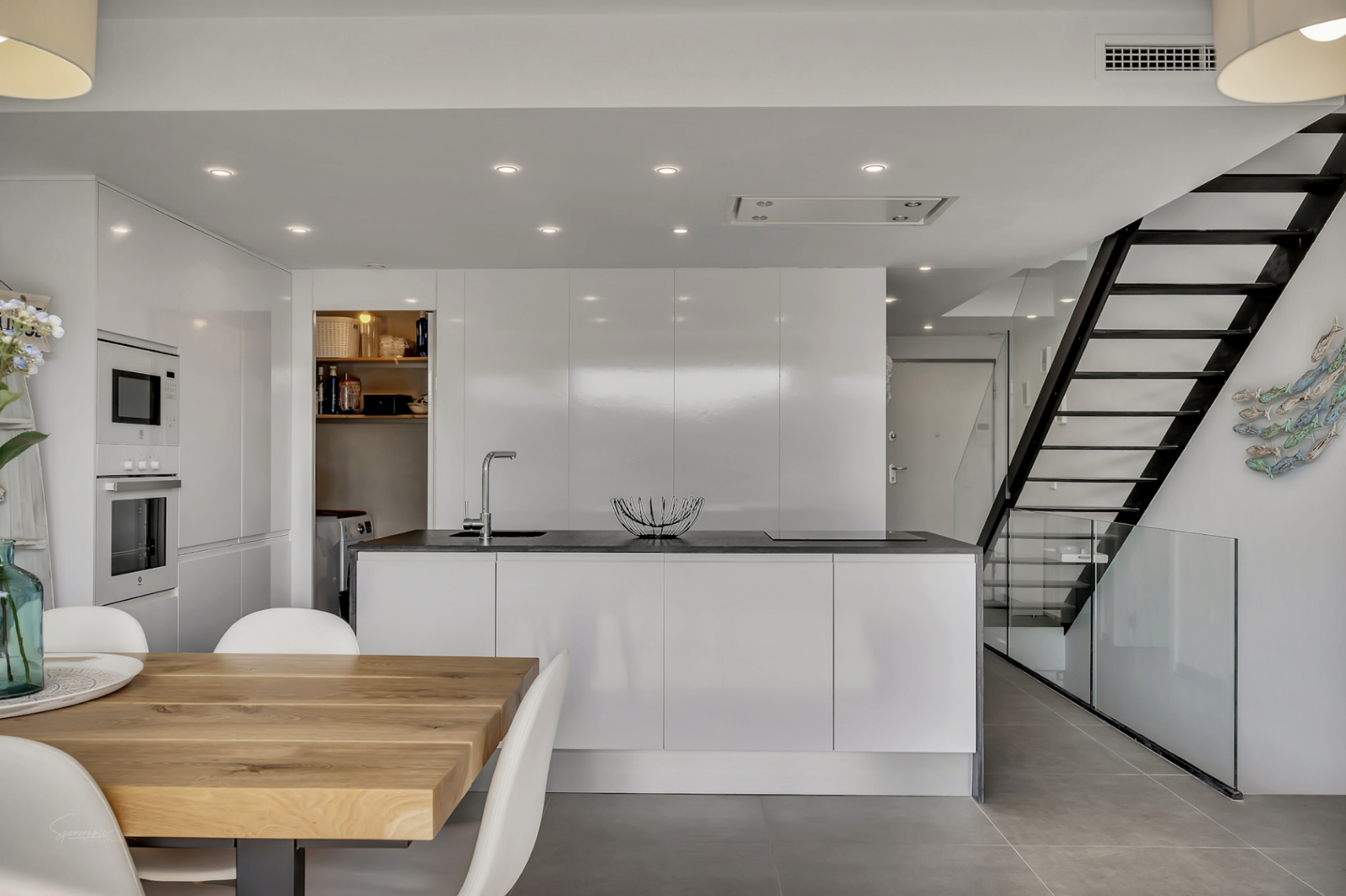 Moderne, offene Küche mit weißer Kochinsel, integrierten Geräten und einer minimalistischen Treppe aus schwarzem Metall und Glas.
