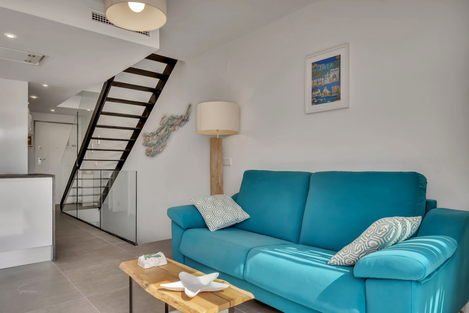 Moderner Wohnbereich mit türkisblauem Sofa, Holz-Couchtisch und einer schwarzen Metalltreppe mit Glasgeländer, die die Ebenen des Triplex verbindet.