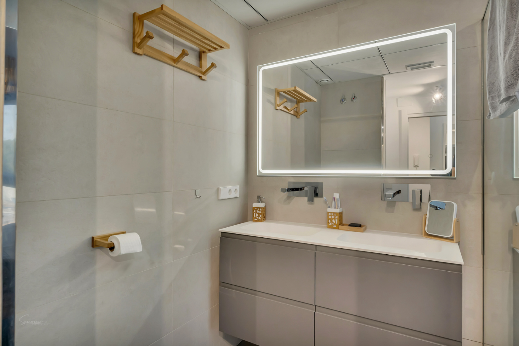 Modernes Badezimmer mit Doppelwaschtisch, grauen Unterschränken und einem großen LED-Spiegel. Holzakzente und neutrale Wandfliesen unterstreichen das elegante Design.
