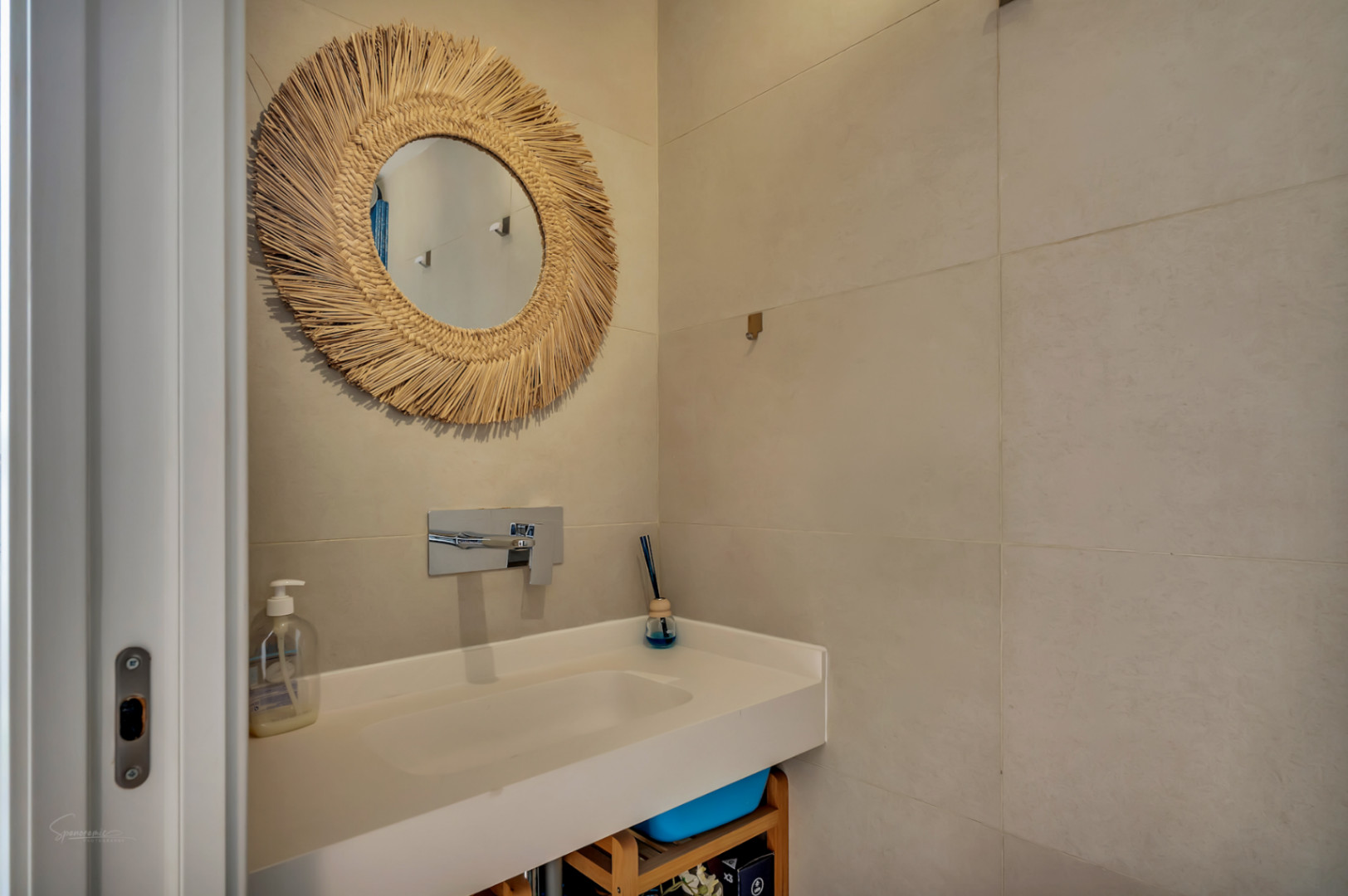 Modernes Badezimmer mit integriertem weißem Waschbecken, Unterputz-Chromarmatur und dekorativem Spiegel mit Naturfaserrahmen vor sandfarbenen großformatigen Wandfliesen.