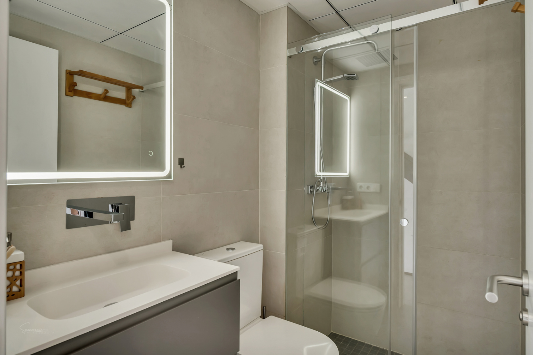 Modernes Badezimmer mit neutralen Fliesen, grauem Unterschrank und LED-Spiegel neben einer begehbaren Dusche mit Glasschiebetür.