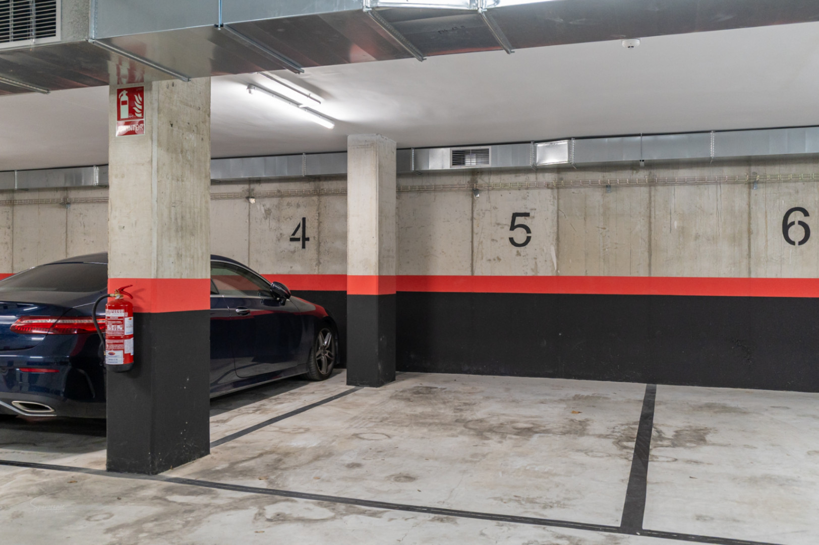 Nummerierter Tiefgaragenstellplatz mit Sichtbetonwänden, industriellen Lüftungsrohren und installierten Brandschutzvorrichtungen.