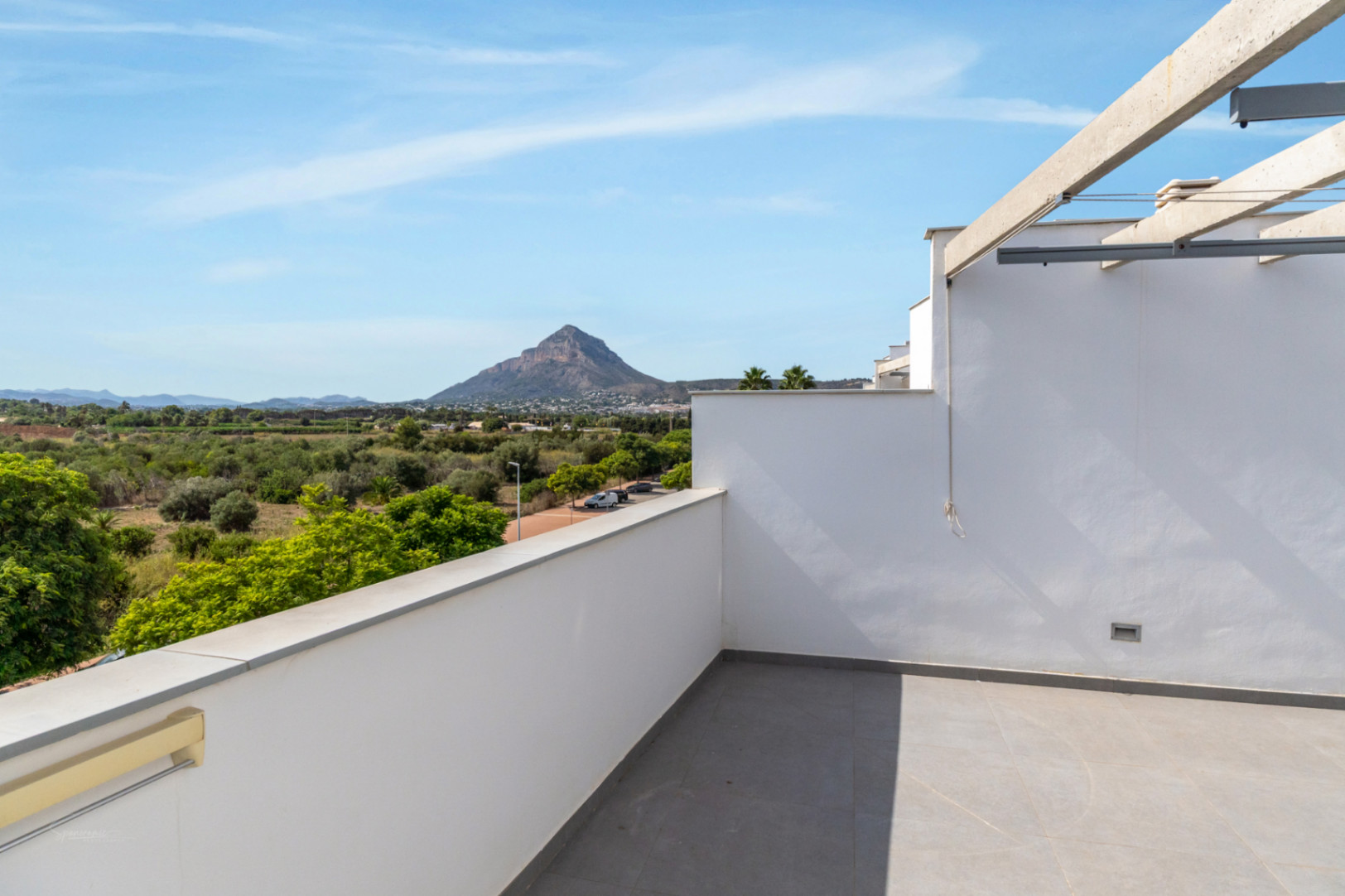 Private Dachterrasse mit grauen Fliesen und Panoramablick auf den Berg Montgó und grüne Felder. Moderne weiße Wände und eine Pergola-Konstruktion aus Beton.