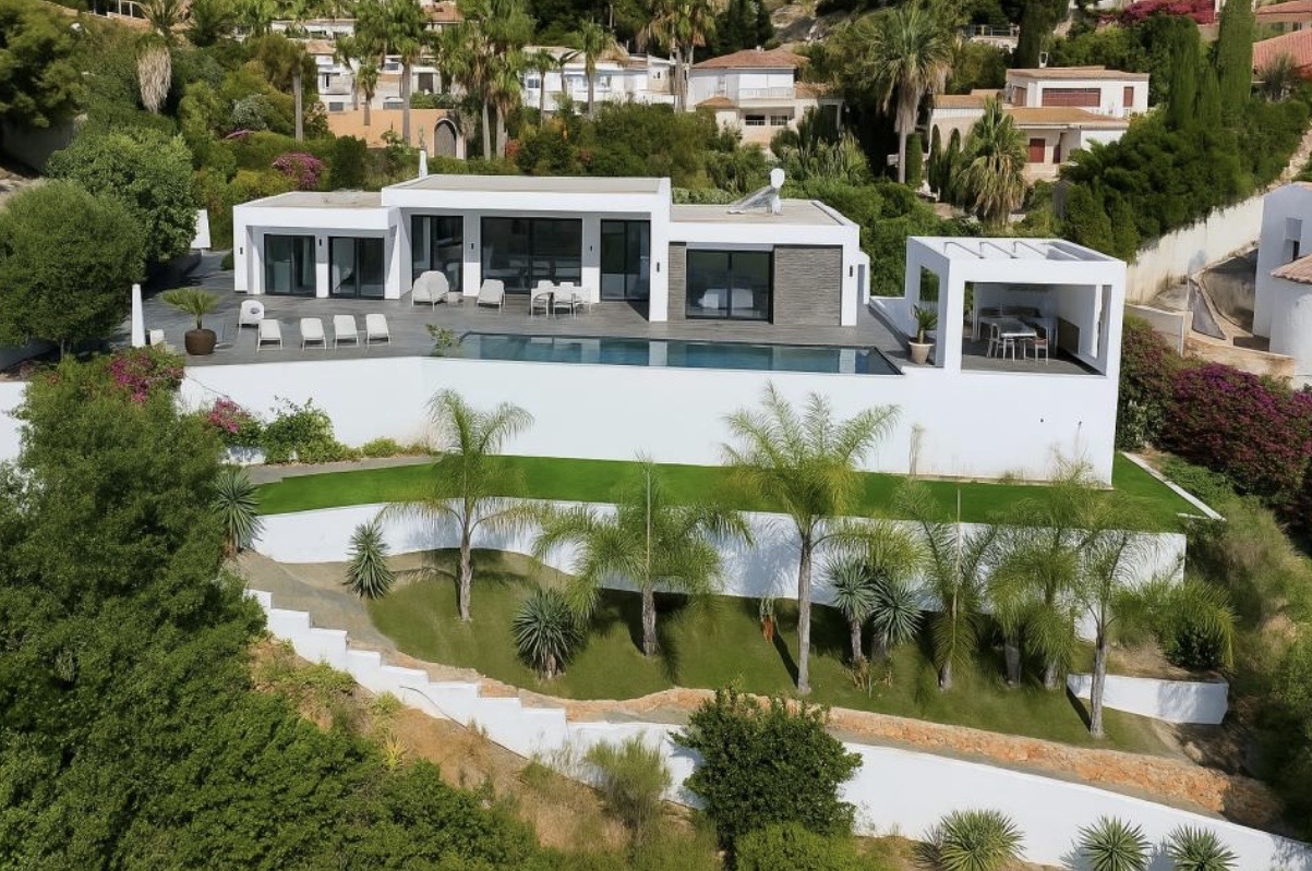 Außenansicht der modernen Villa mit großer Terrasse, Infinity-Pool und terrassiertem mediterranem Garten mit Palmen.