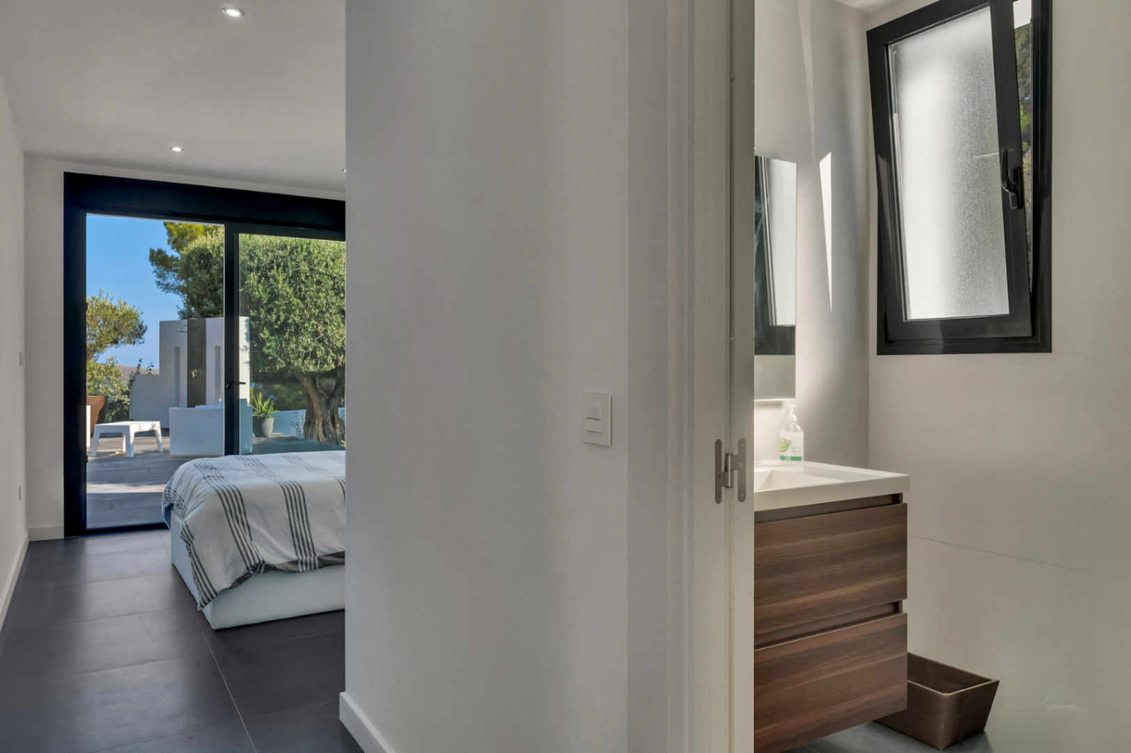 En-Suite-Schlafzimmer mit dunklem Holzwaschtisch und direktem Terrassenzugang. Bodentiefe Schiebefenster geben den Blick auf die Außendusche und den Garten frei.
