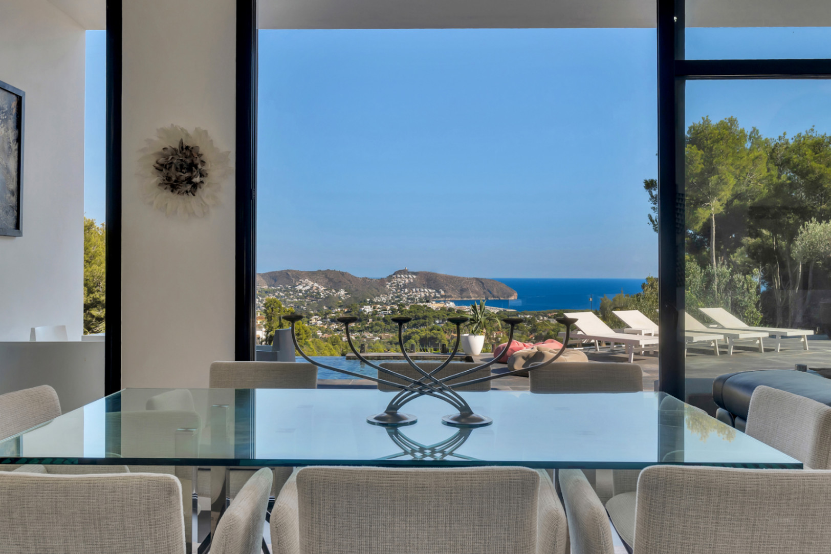 Essbereich mit Glastisch und gepolsterten Stühlen, der durch bodentiefe Fenster einen weiten Panoramablick auf das Meer und die Küste bietet.