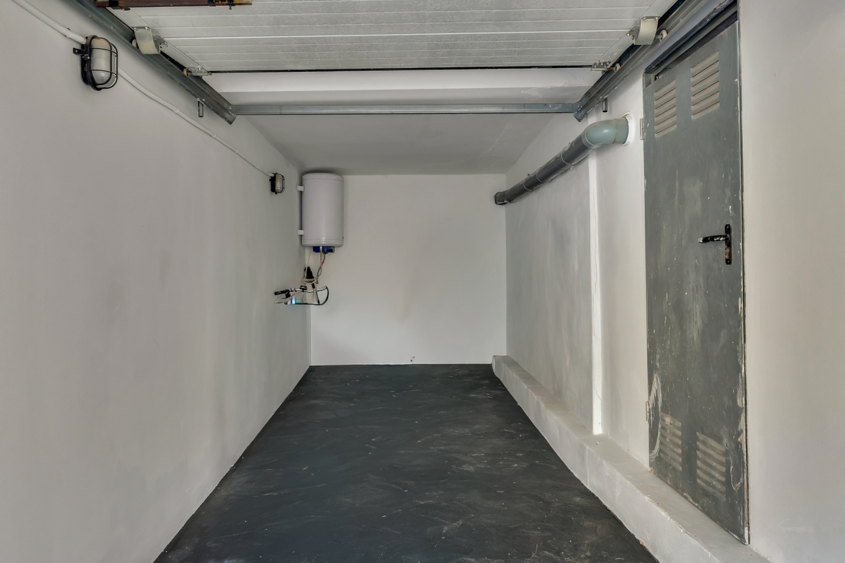Funktionale Garage mit dunklem Boden, weißen Wänden, elektrischem Warmwasserbereiter und seitlicher Metalltür für zusätzlichen Stauraum.