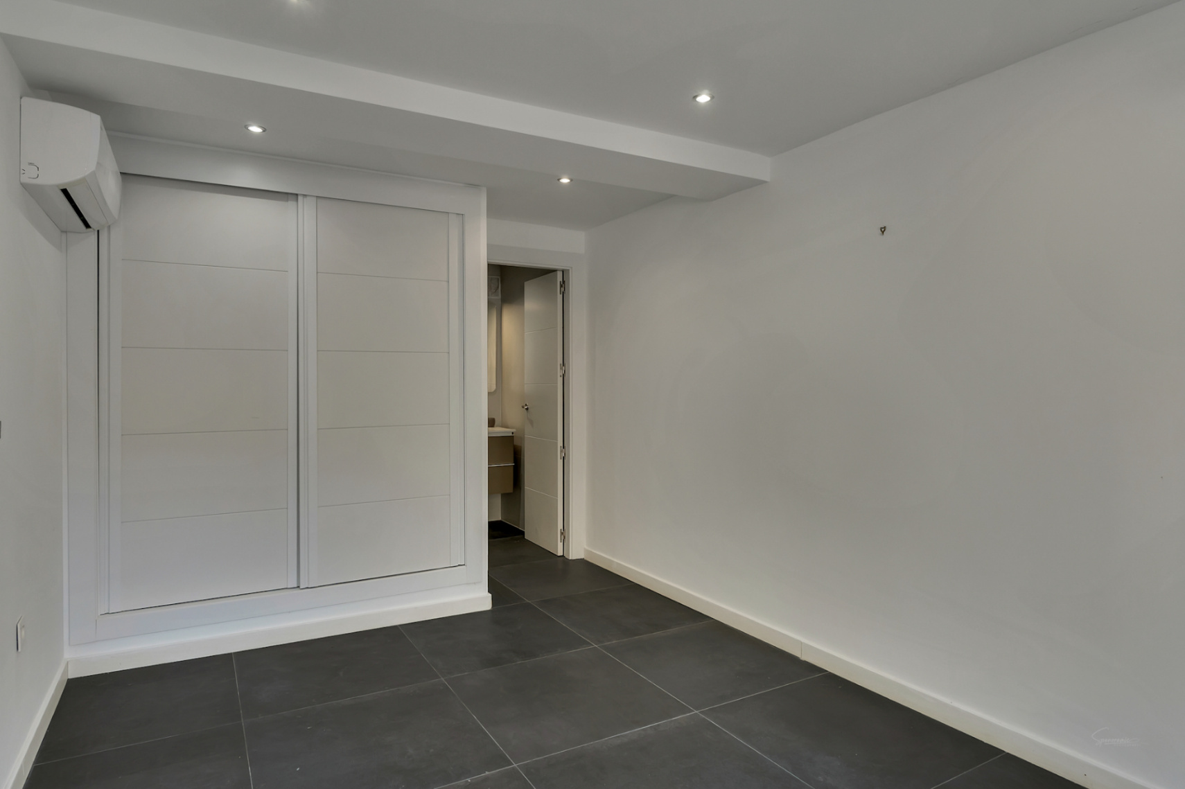 Funktionales Schlafzimmer mit weißem Einbauschrank, dunkelgrauen Bodenfliesen, Klimaanlage und Zugang zum privaten En-Suite-Badezimmer.