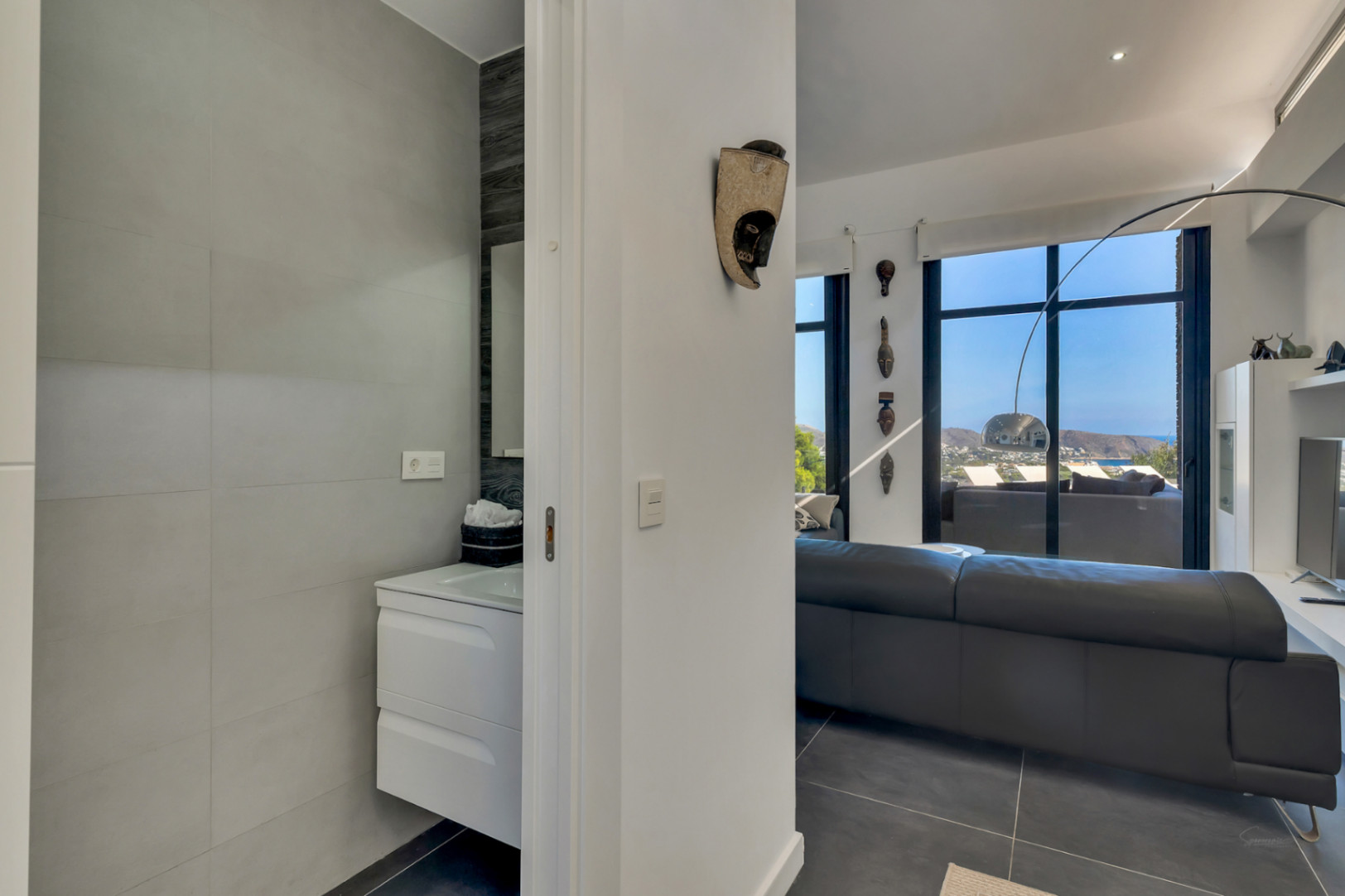 Gäste-WC mit hängendem Waschtisch neben dem Wohnzimmer, das durch große Fensterfronten einen weiten Blick auf das Meer und die Küste freigibt.