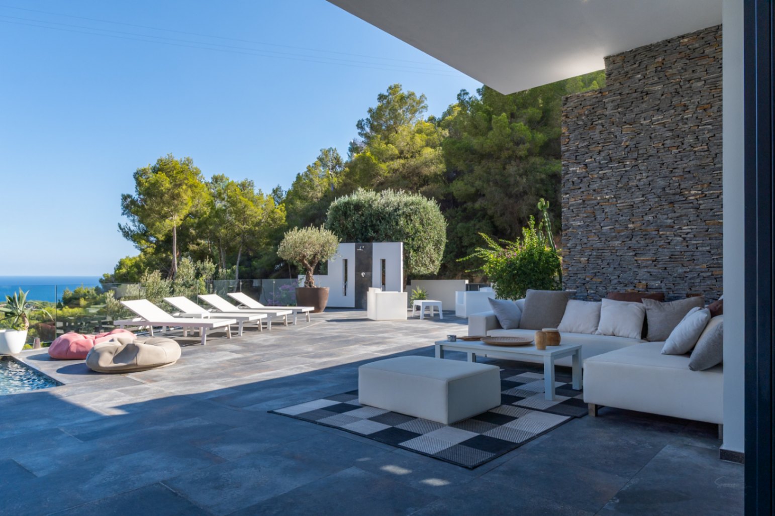 Großzügige Terrasse mit weißer Lounge-Ecke vor einer Natursteinwand, angrenzendem Sonnendeck mit Liegen und weitem Blick auf das Mittelmeer.