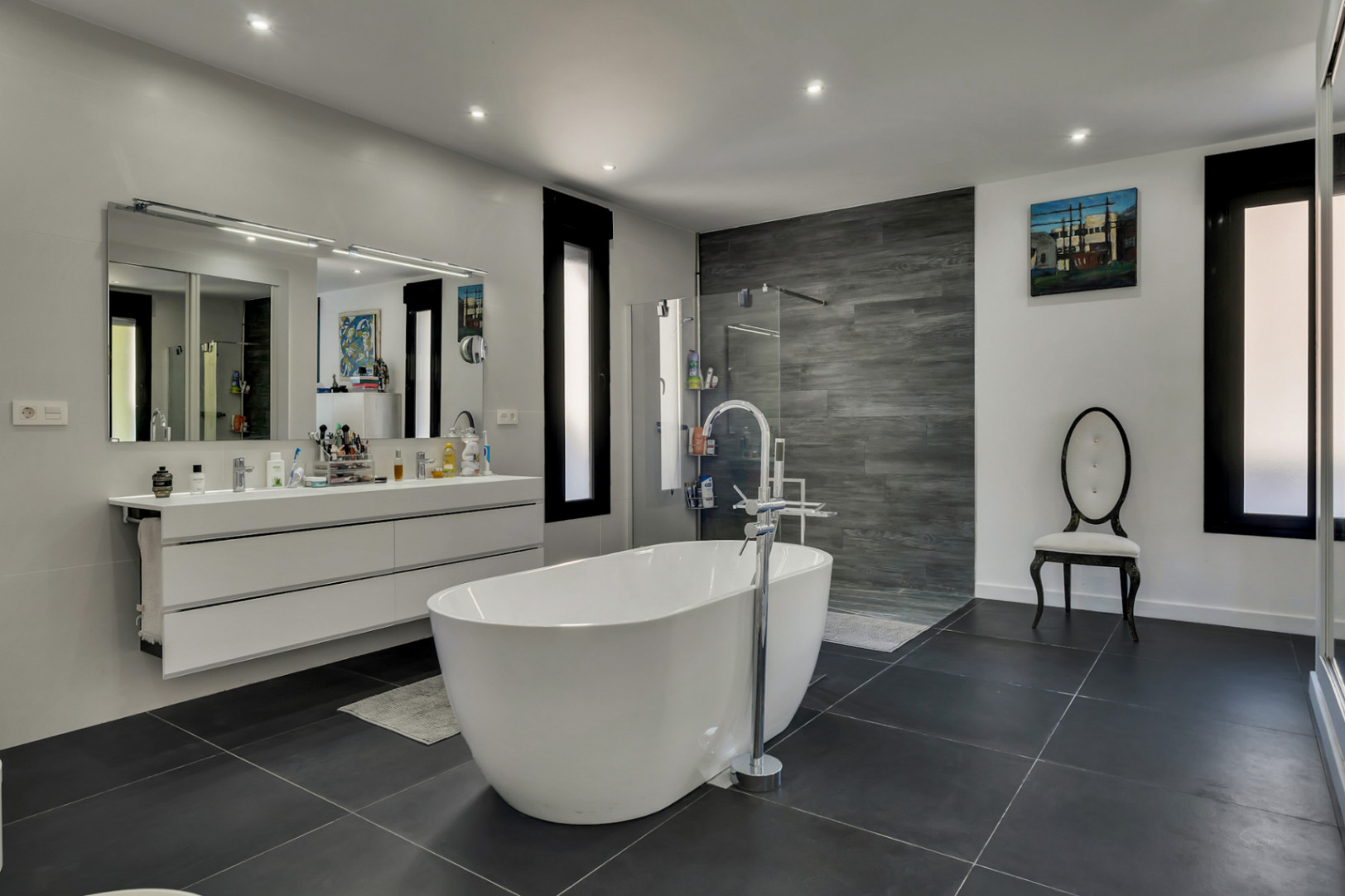 Hauptbadezimmer mit freistehender Badewanne, Doppelwaschtisch und begehbarer Dusche mit grauer Holzoptik-Wand und dunklen Bodenfliesen.