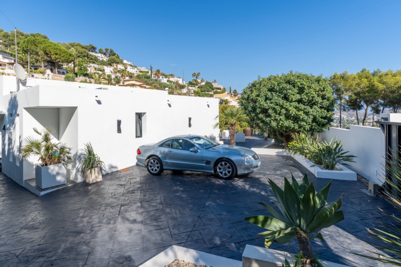 Haupteingang mit Parkplatz aus dunklem Stempelbeton, minimalistischer weißer Fassade und integrierten Pflanzgefäßen mit mediterraner Bepflanzung.