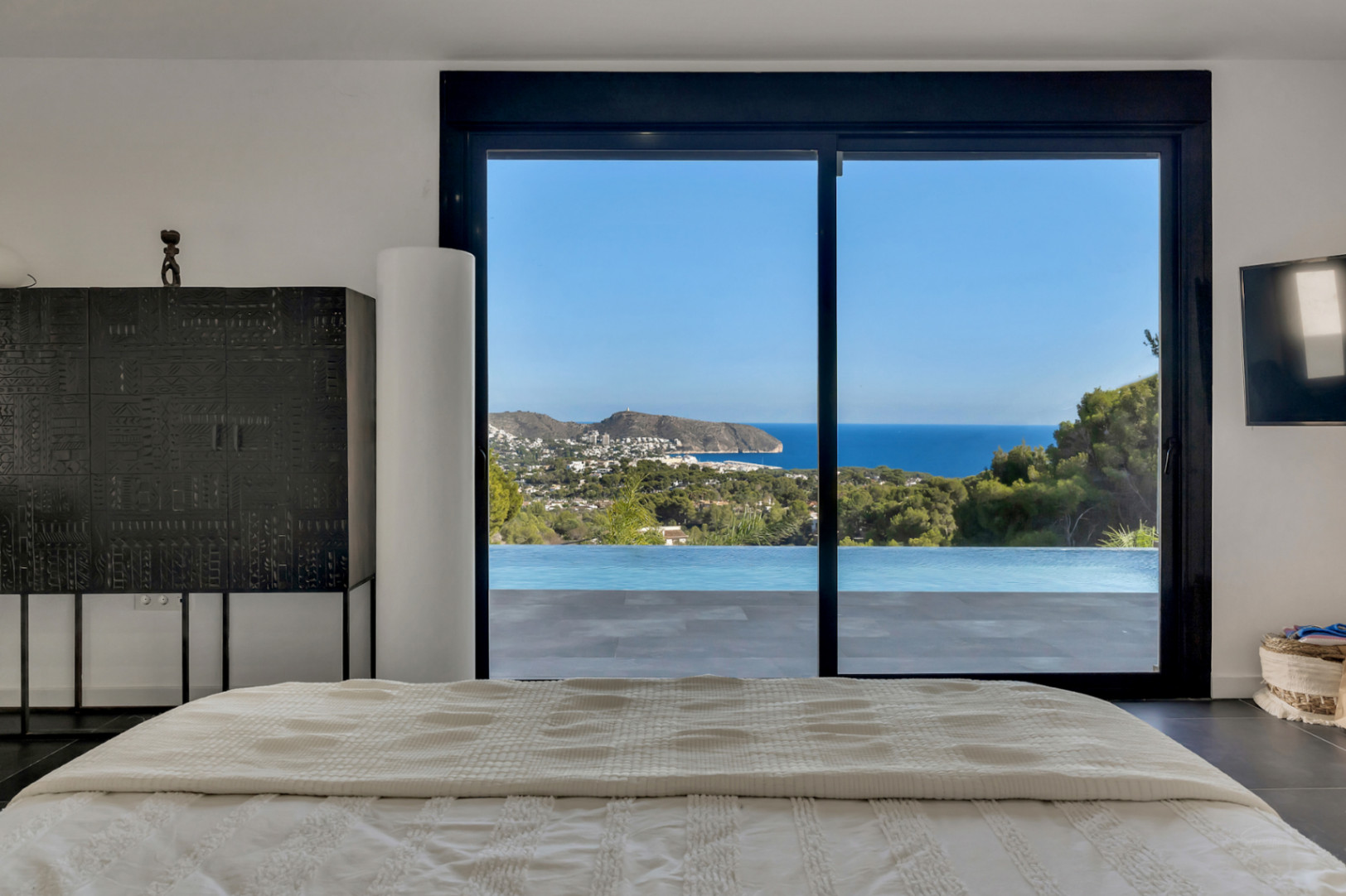 Hauptschlafzimmer mit Panoramablick auf das Meer und den Infinity-Pool durch große Glasschiebetüren. Mit Designermöbeln und dunklen Bodenfliesen.