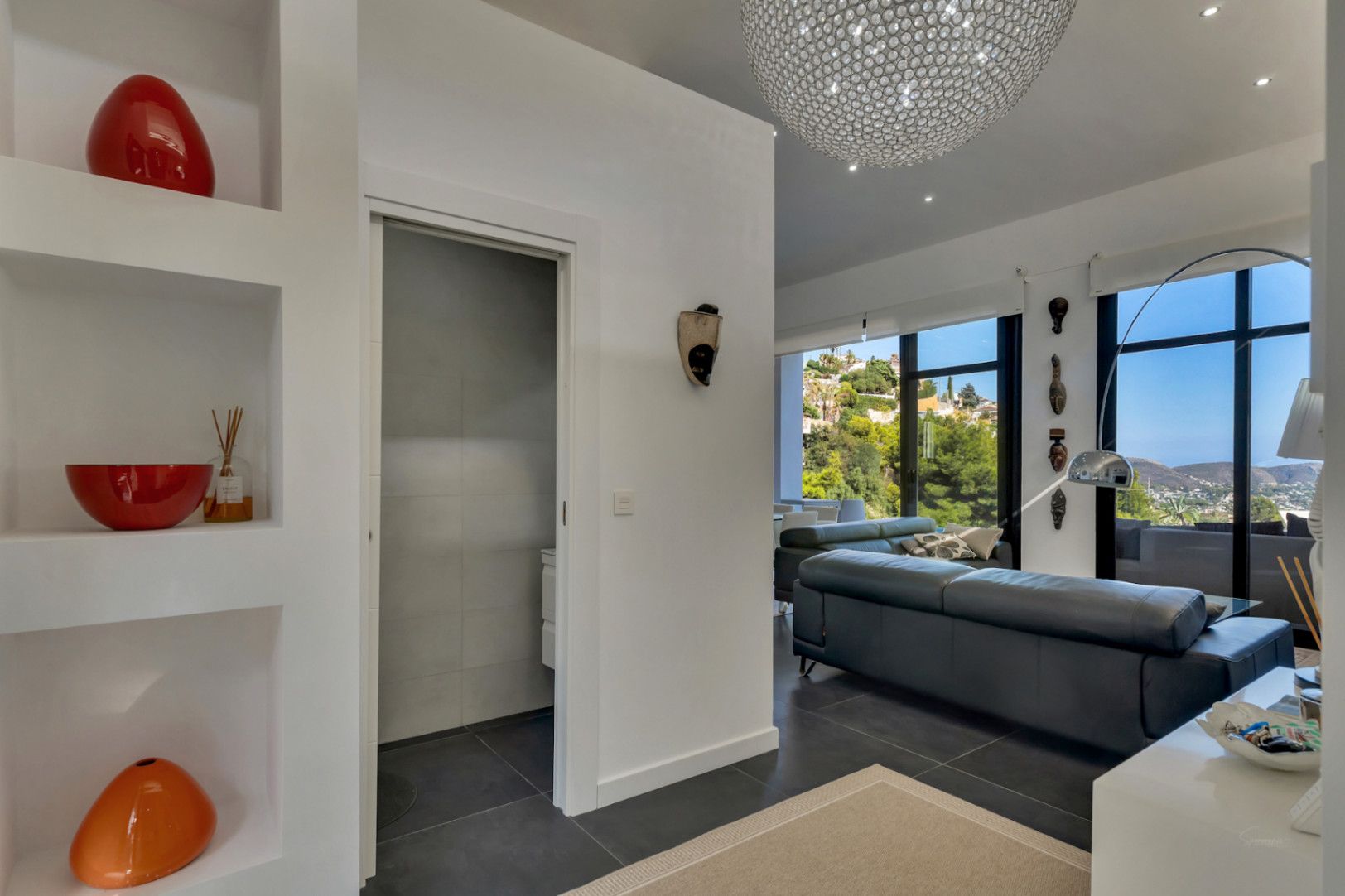 Heller Eingangsbereich mit integrierten Wandnischen, dunklem Fliesenboden und Blick in das Wohnzimmer mit großen Fensterfronten und Meerfernsicht.