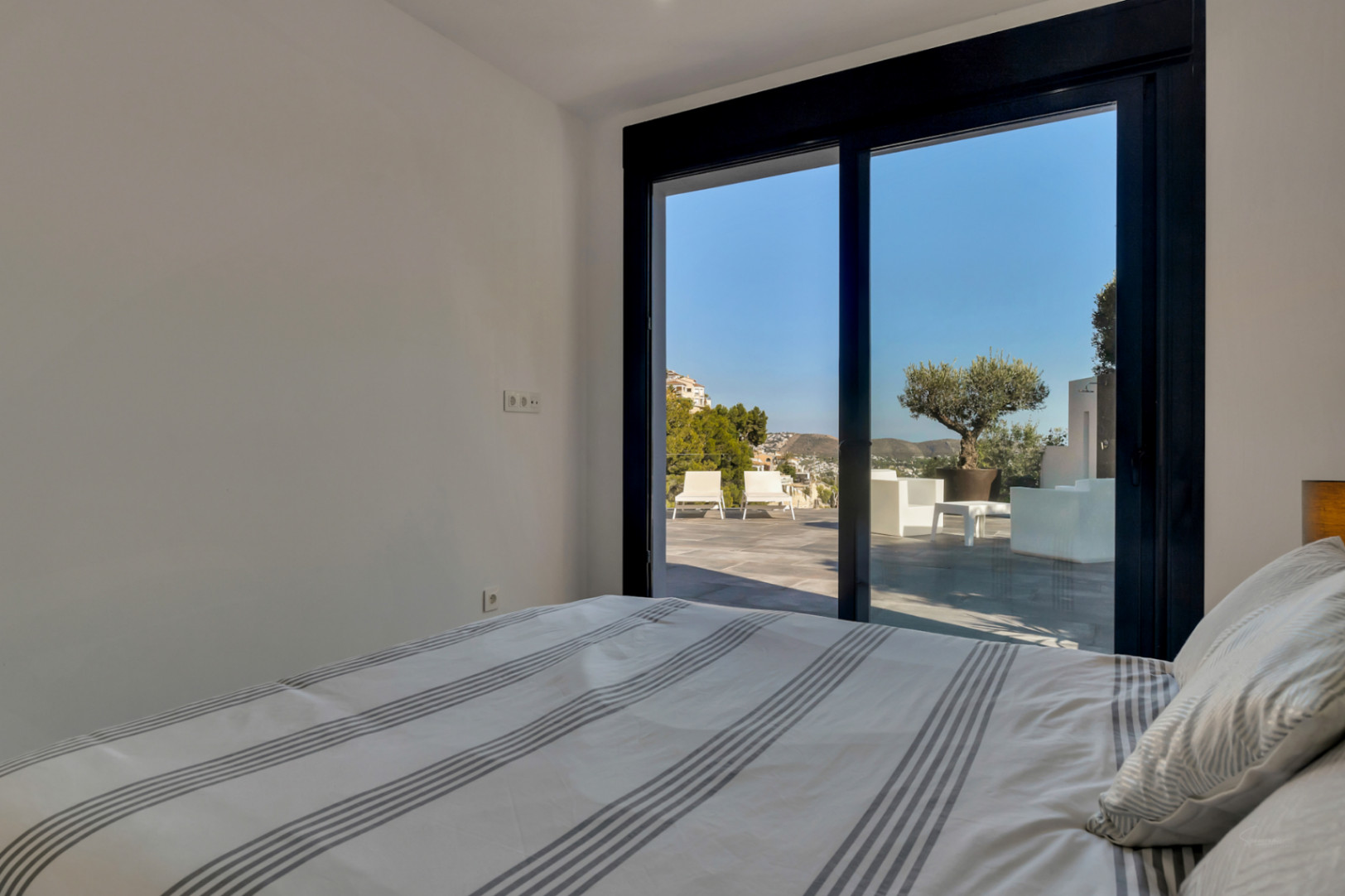 Helles Schlafzimmer mit direktem Terrassenzugang durch große Glasschiebetüren. Der Blick reicht über die Lounge-Möbel und einen Olivenbaum bis zu den Hügeln.