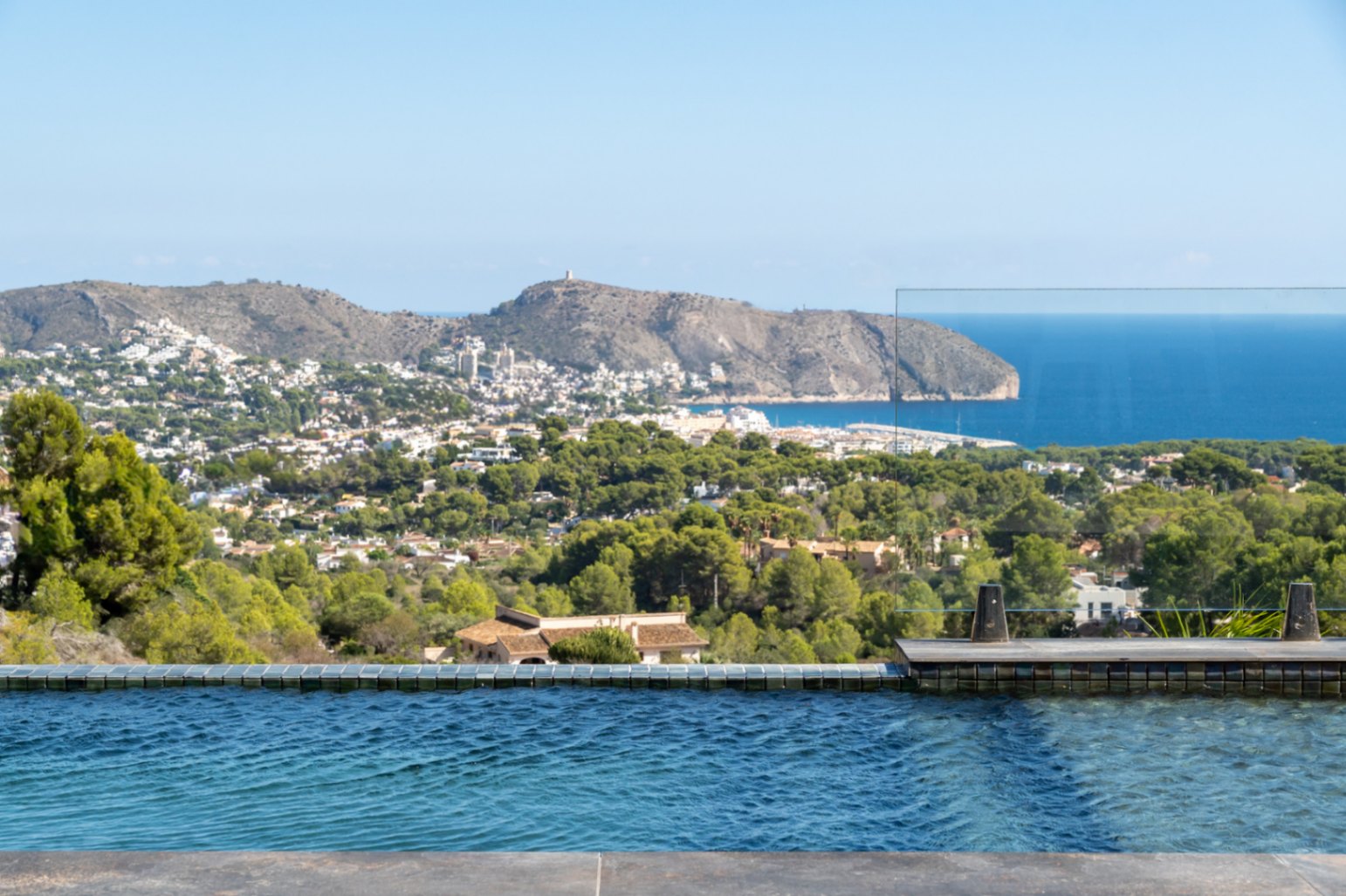 Infinity-Pool mit blauen Mosaikfliesen und Glasgeländer, der einen Panoramablick auf das Mittelmeer, das Cap d'Or und die grüne Küstenlandschaft bietet.