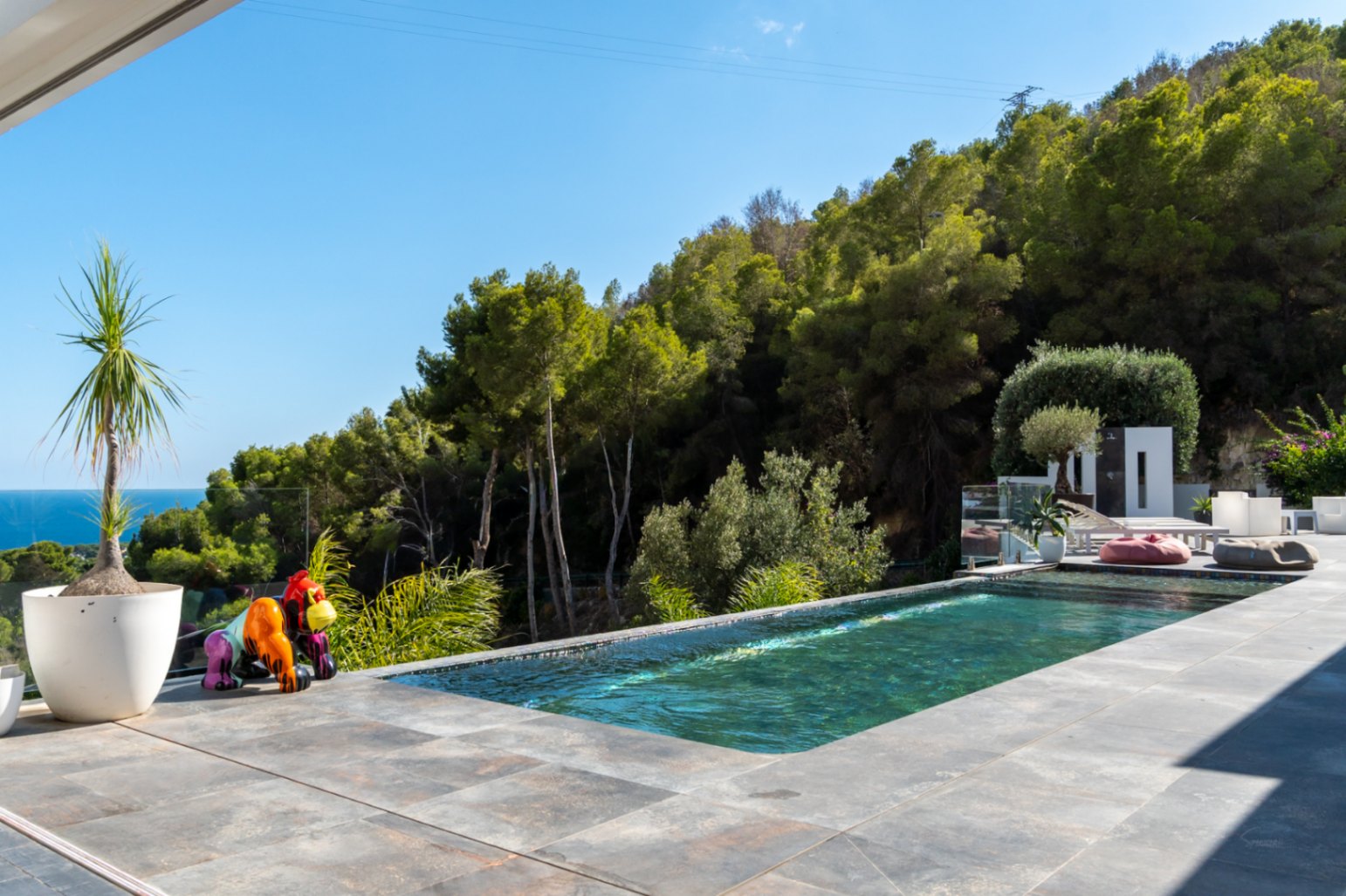 Infinity-Pool mit großformatiger Fliesenterrasse, Blick auf das Meer und den Pinienwald, ergänzt durch eine farbenfrohe Gorilla-Skulptur.