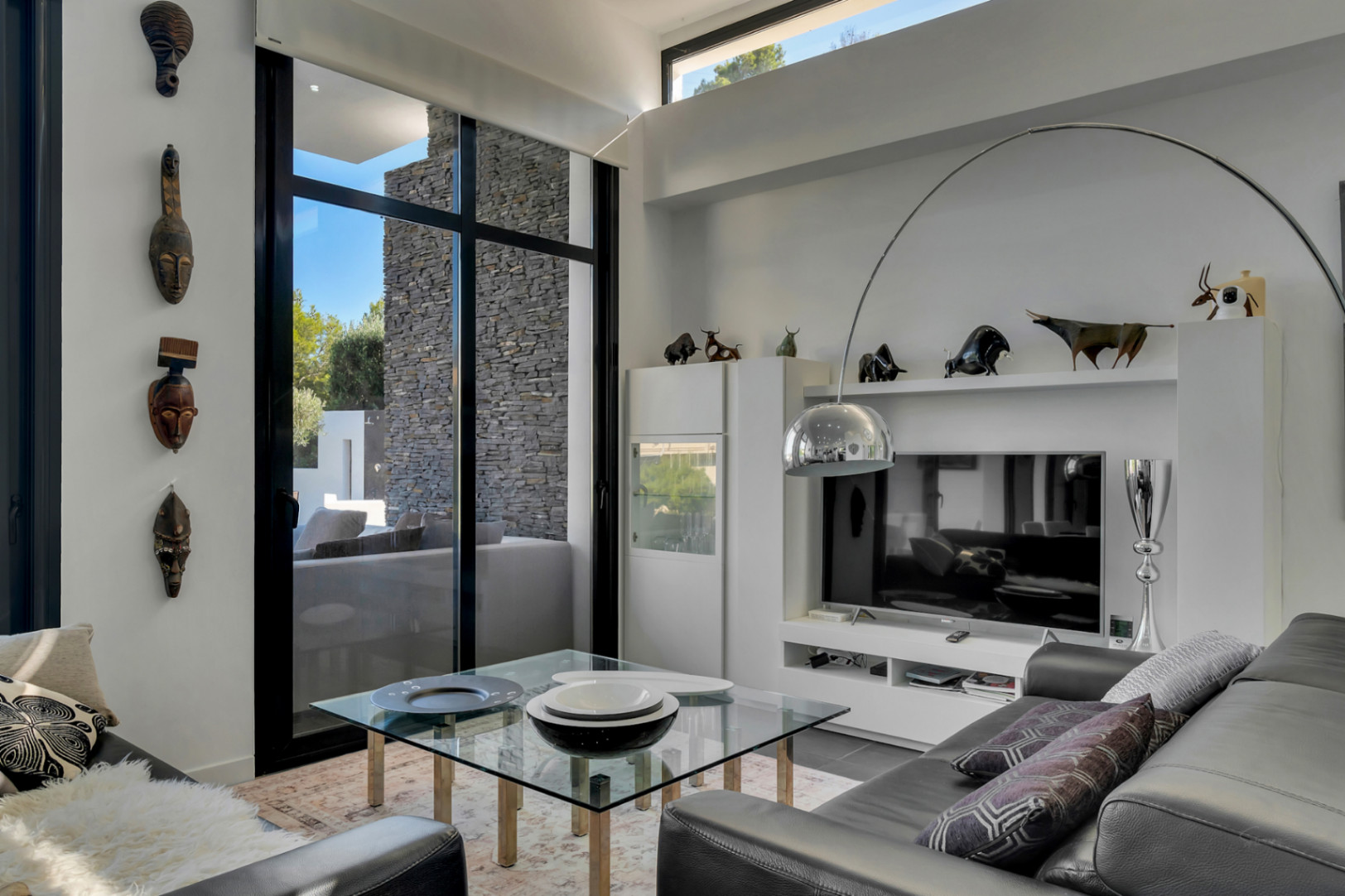 Moderner Wohnraum mit hohen Decken, schwarzen Fensterprofilen und ethnischer Kunst. Ausgestattet mit einer Bogenlampe aus Chrom und Blick auf die Natursteinwand im Außenbereich.