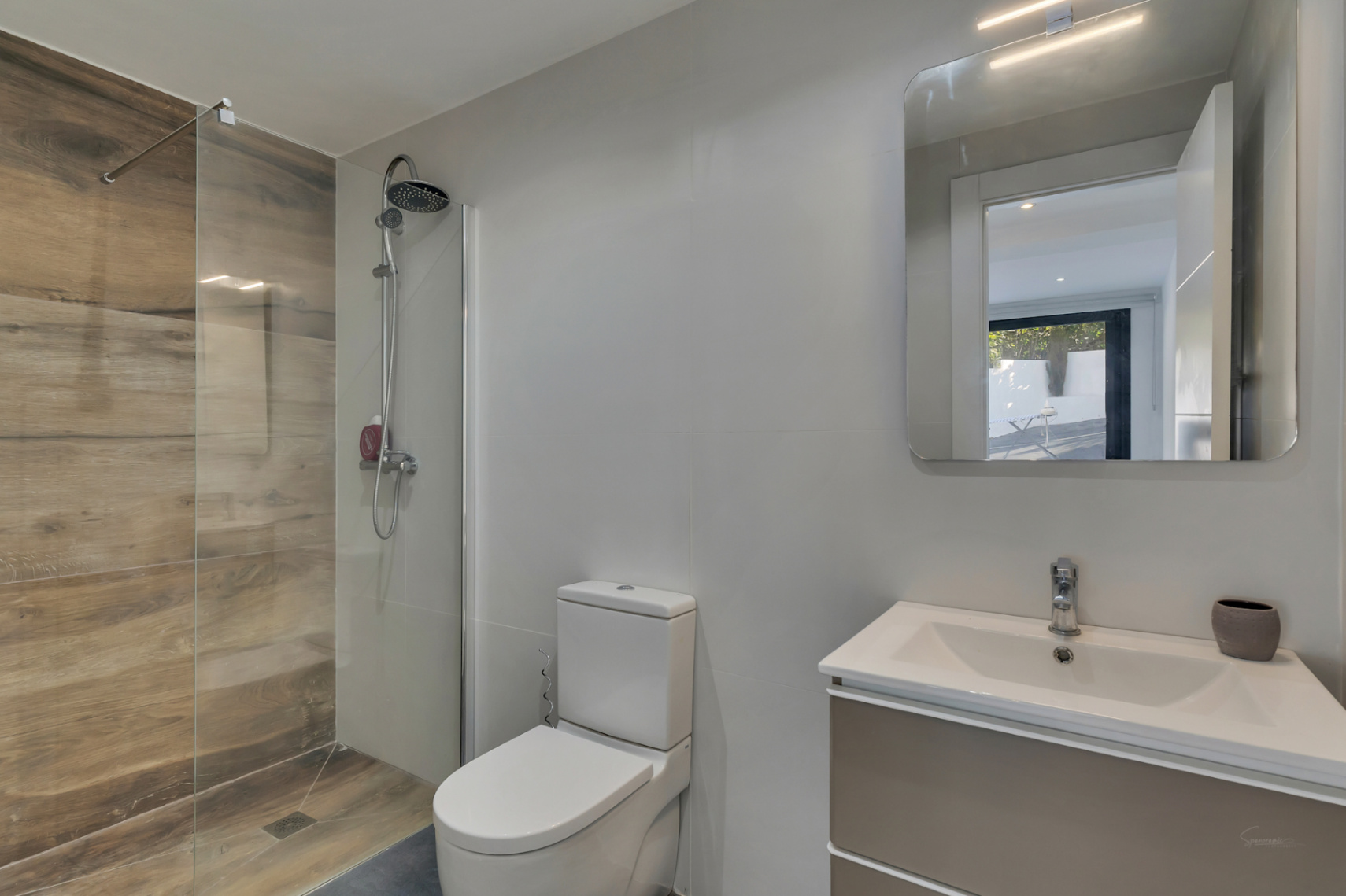 Modernes Badezimmer mit begehbarer Dusche in Holzoptik-Fliesen, fester Glaswand und hängendem Waschtisch mit beleuchtetem Spiegel.