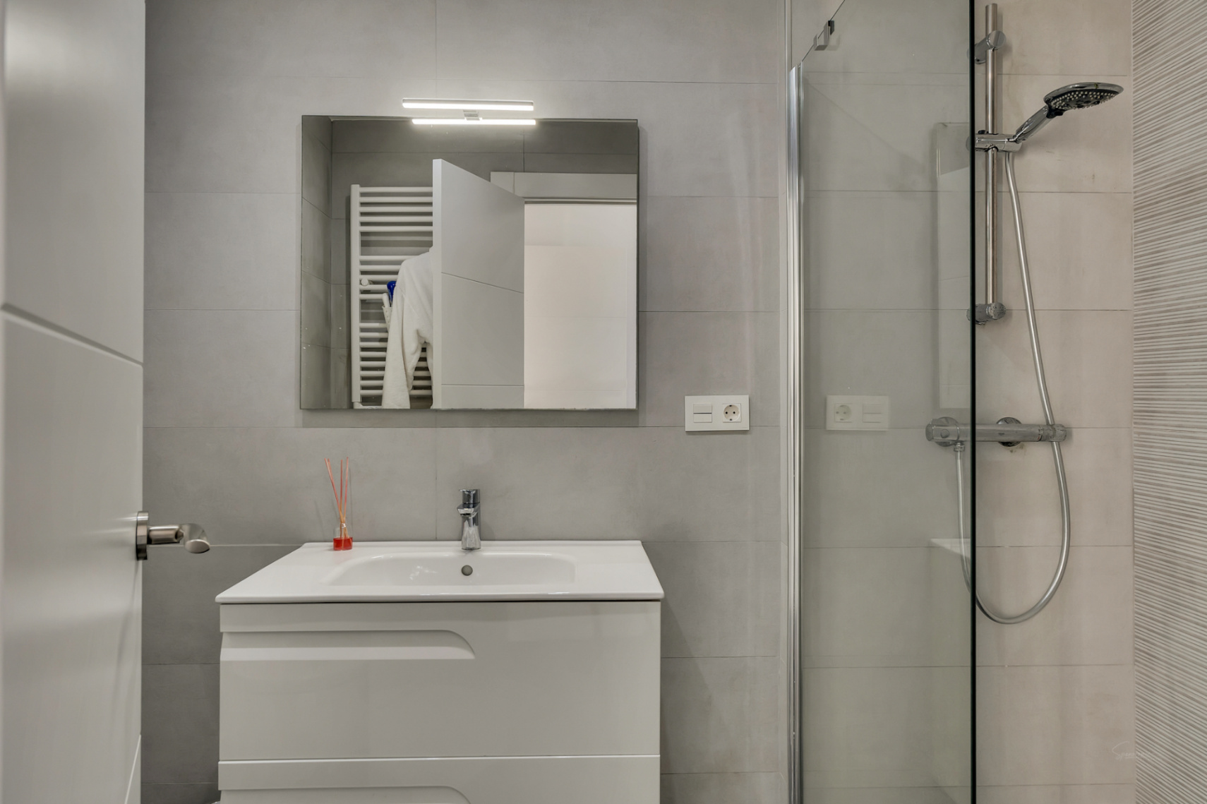 Modernes Badezimmer mit weißem Waschtisch, beleuchtetem Spiegel und einer begehbaren Dusche mit Glastrennwand und grauen Fliesen.