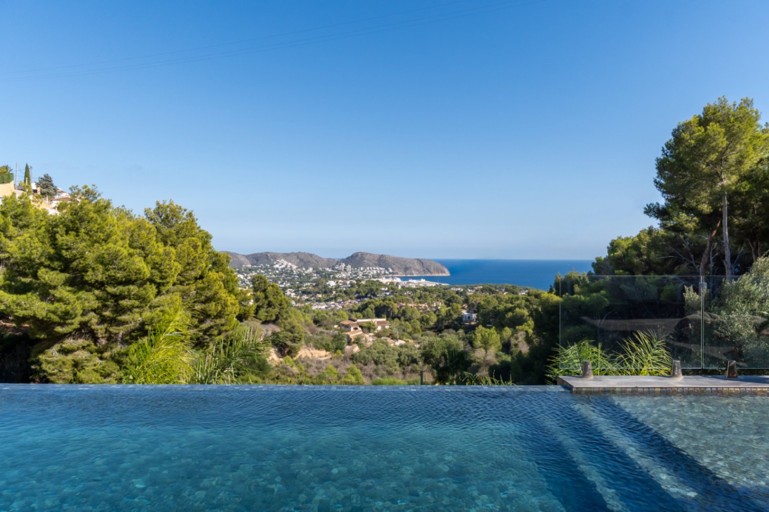 Panoramablick auf das Mittelmeer und die Küste von Moraira vom Infinity-Pool aus, umgeben von üppigen Pinien und mediterraner Vegetation.