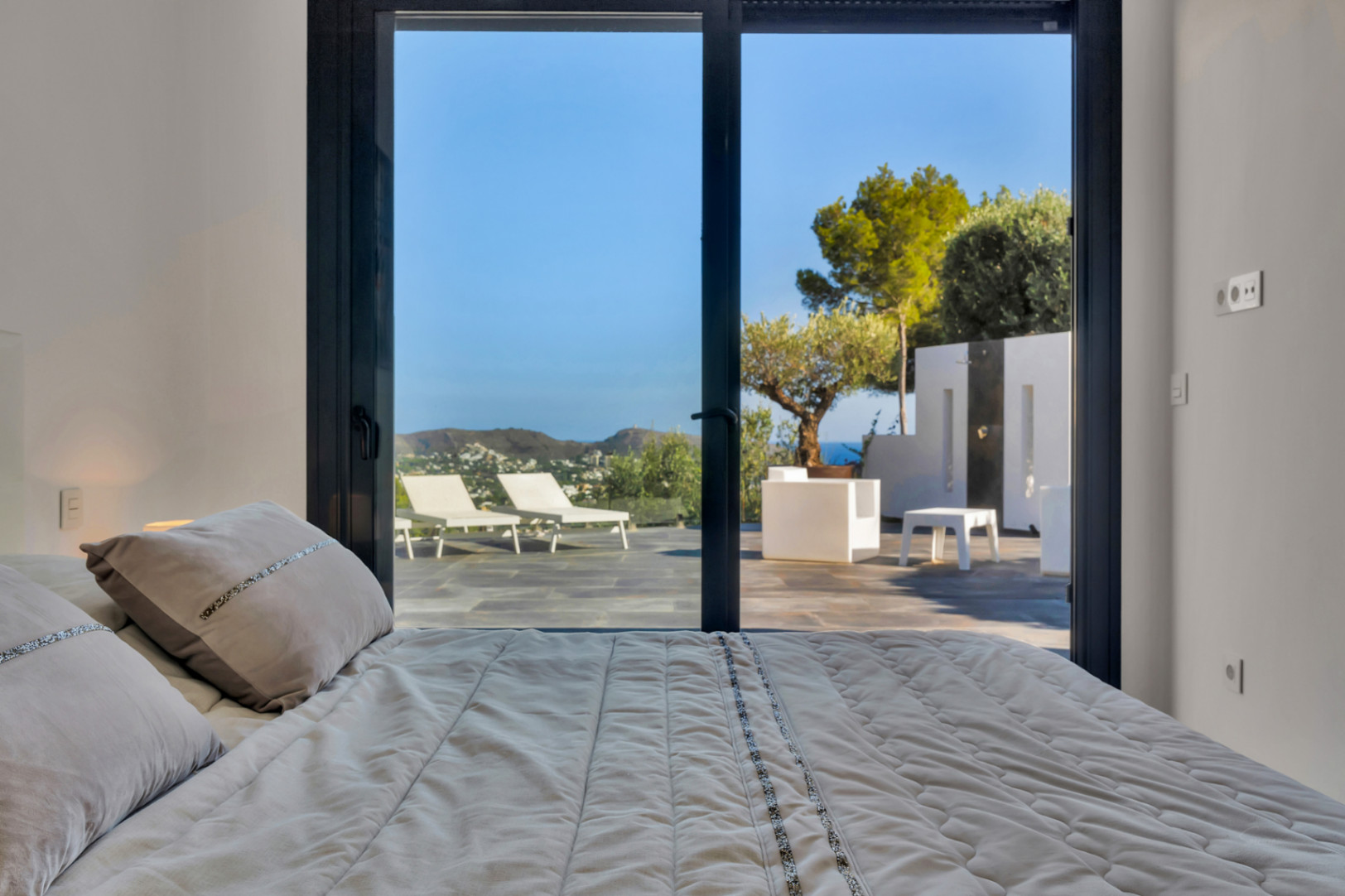 Schlafzimmer mit großen Glasschiebetüren, die direkt auf die Steinterrasse mit Sonnenliegen, Außendusche und Panoramablick auf das Meer führen.