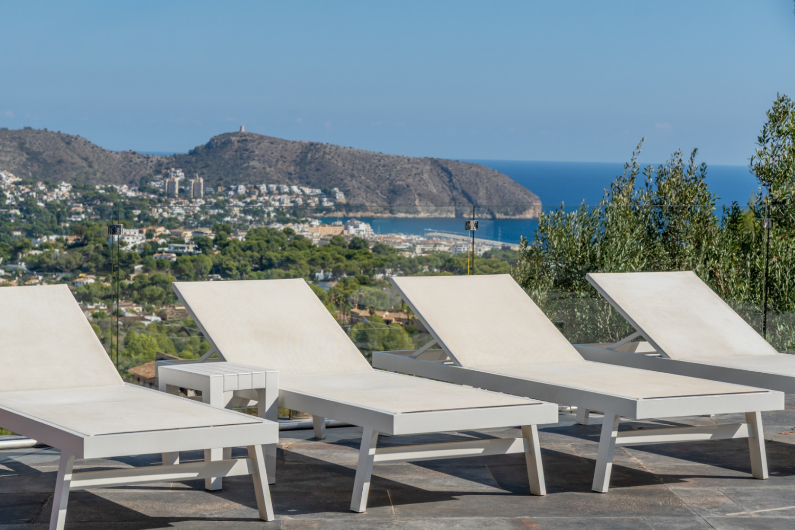 Sonnenterrasse mit modernen weißen Liegen und Glasgeländer, die einen Panoramablick auf das Mittelmeer und die Bucht von Moraira bietet.