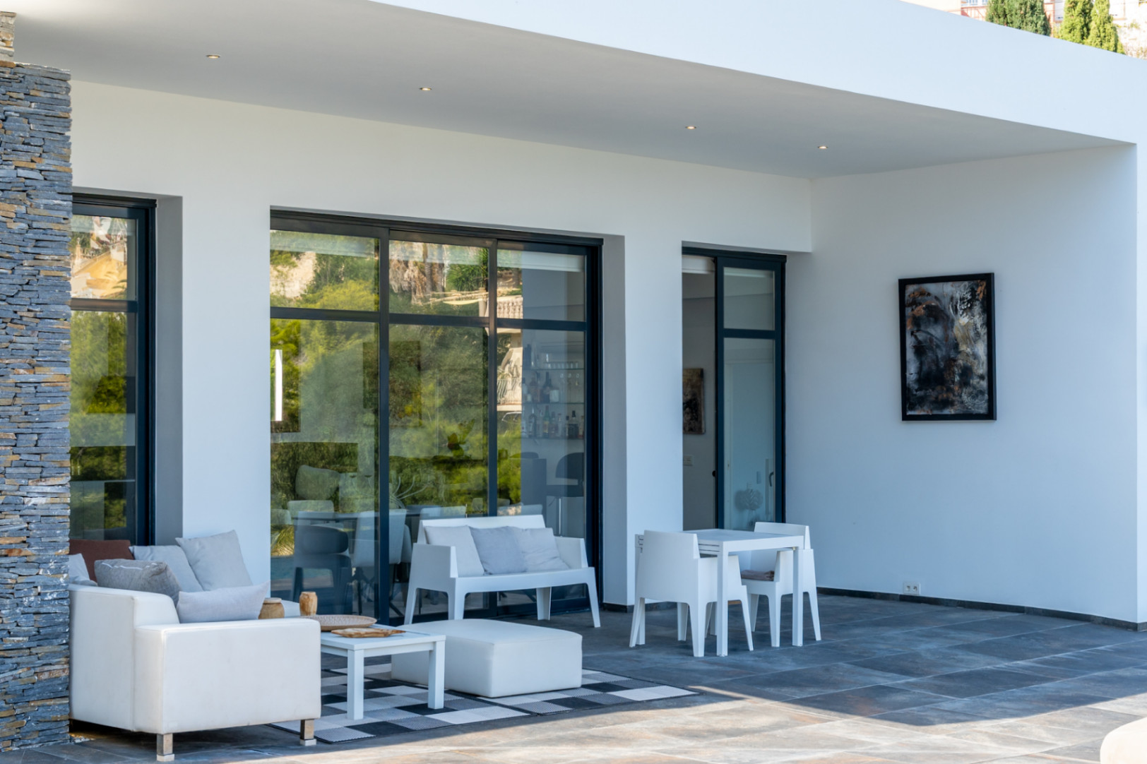 Überdachte Terrasse mit dunklem Feinsteinzeugboden, Natursteinverkleidung und minimalistischen weißen Außenmöbeln vor großen Schiebefenstern.