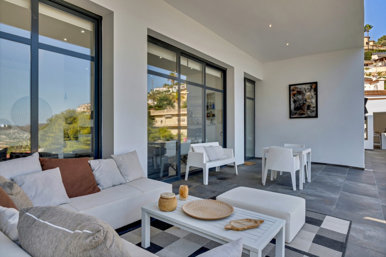 Überdachte Terrasse mit modernem Loungebereich, grauen Bodenfliesen und großen Glasfronten, die die Umgebung widerspiegeln.