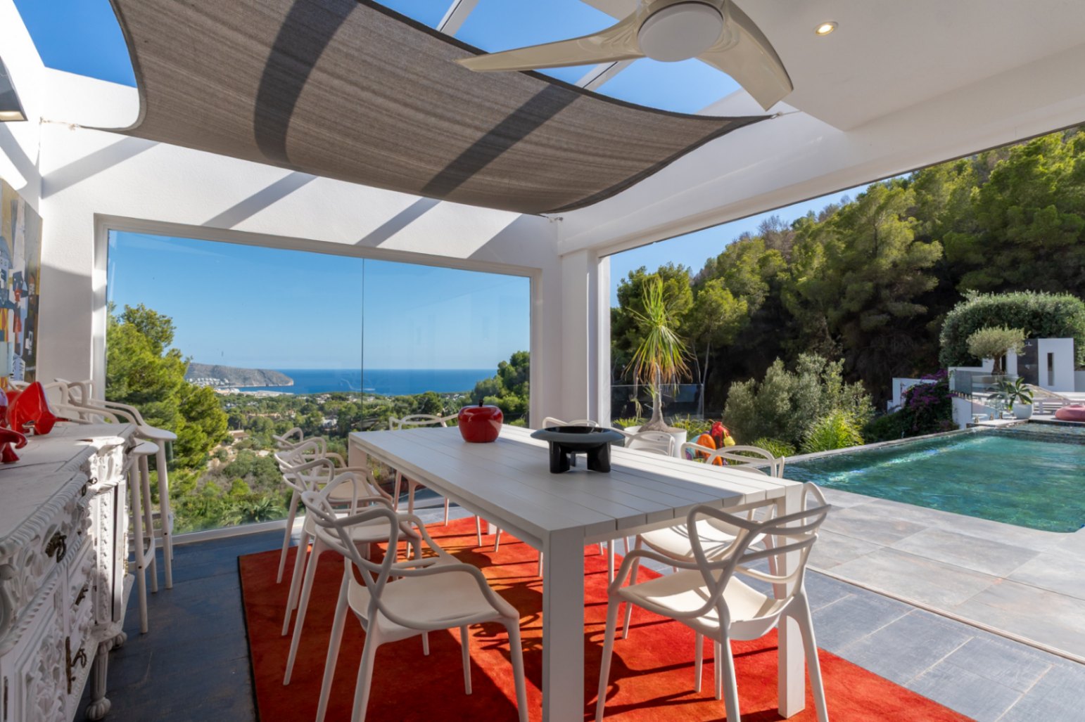 Verglaster Außen-Essbereich mit Panoramablick auf das Meer, Designermöbeln, Sonnensegel und direktem Zugang zum Infinity-Pool inmitten mediterraner Natur.