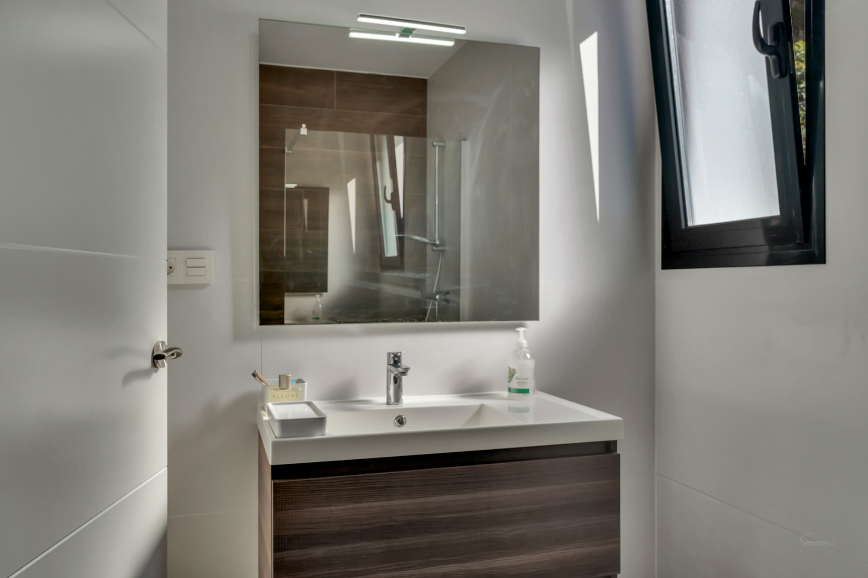 Zeitgenössisches Badezimmer mit schwebendem Waschtisch in dunkler Holzoptik, weißem Becken und beleuchtetem Spiegel. Schwarzes Fenster für Belüftung.