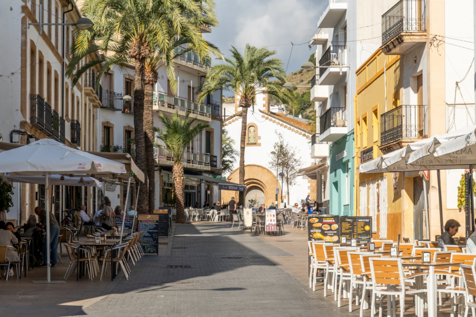 Fußgängerzone in der Altstadt von Jávea mit Außenterrassen, Palmen und traditionellen Gebäuden, die zu einer historischen Kapelle führen.
