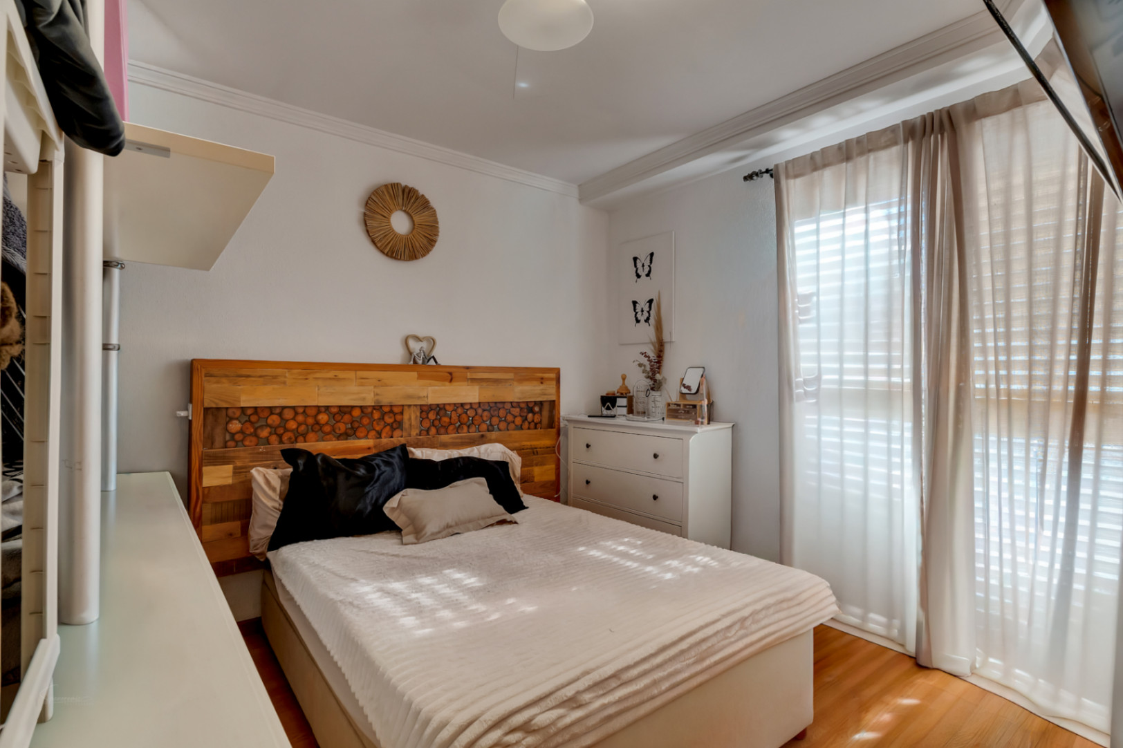 Gemütliches Schlafzimmer mit Doppelbett, handgefertigtem Holzkopfteil und Laminatboden. Ausgestattet mit weißer Kommode und großem Fenster mit lichtdurchlässigen Vorhängen.