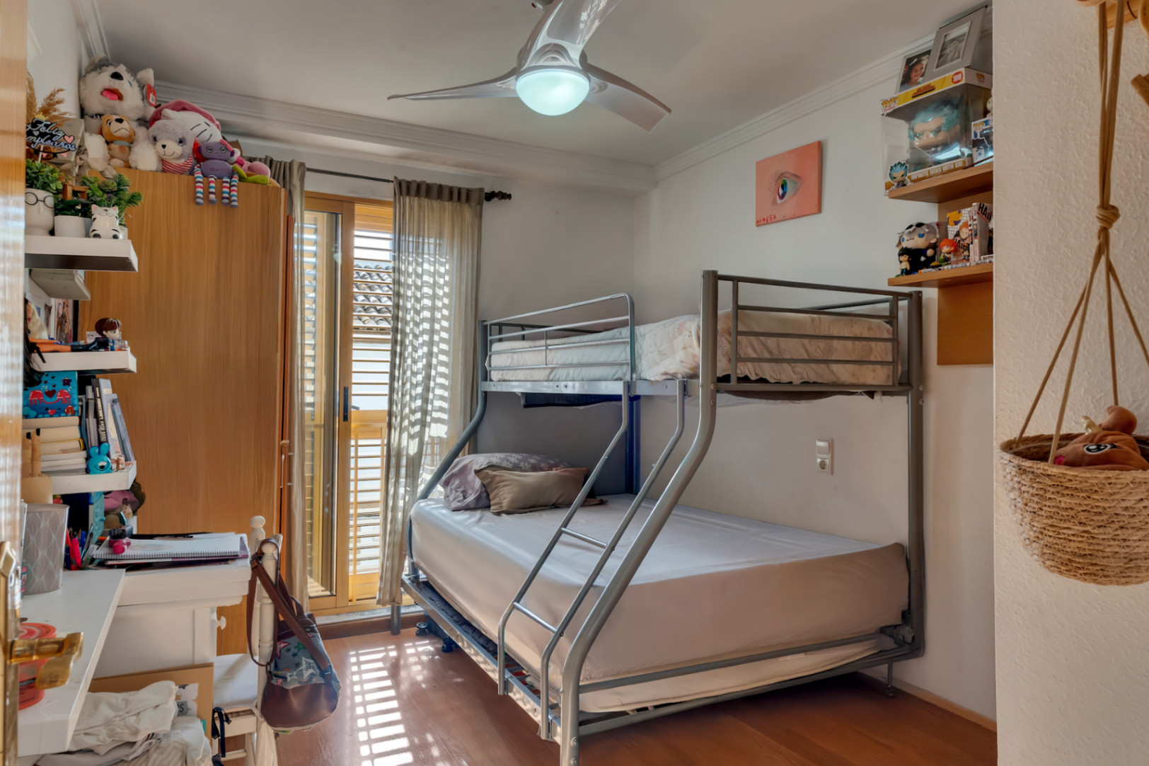 Schlafzimmer mit Metall-Etagenbett, Deckenventilator und Parkettboden. Große Glastüren mit traditionellen Fensterläden führen auf einen lichtdurchfluteten Balkon.