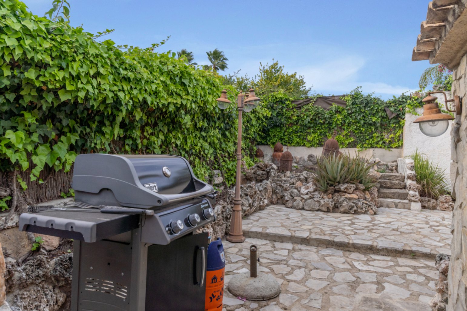 Außenbereich mit Grillstation, unregelmäßiger Steinpflasterung und efeubewachsenen Mauern, ergänzt durch einen Steingarten und rustikale Außenbeleuchtung.