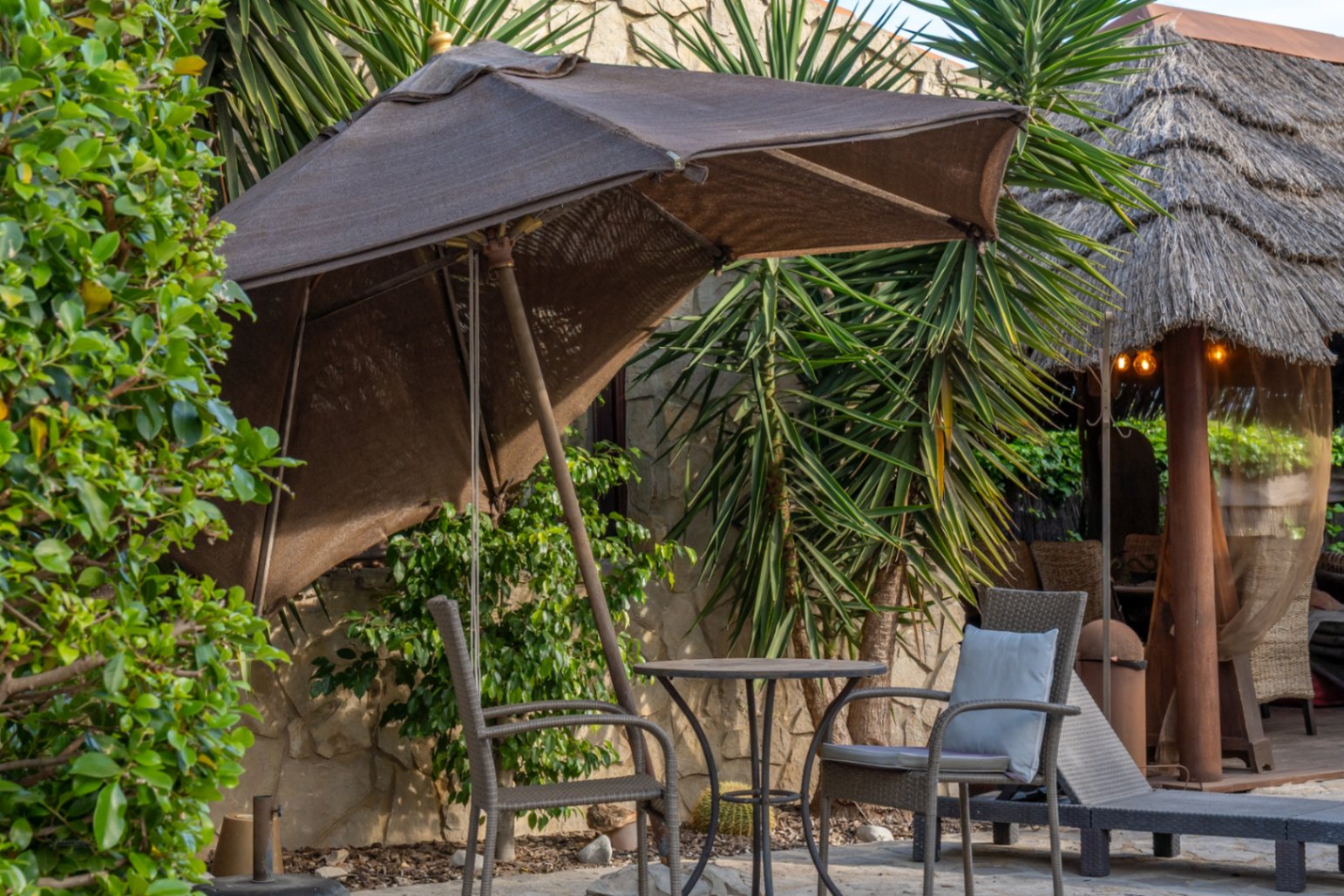Außenbereich mit Rattansesseln, Bistrotisch und Sonnenschirm, angrenzend an eine balinesische Pergola mit Reetdach und Natursteinwänden.