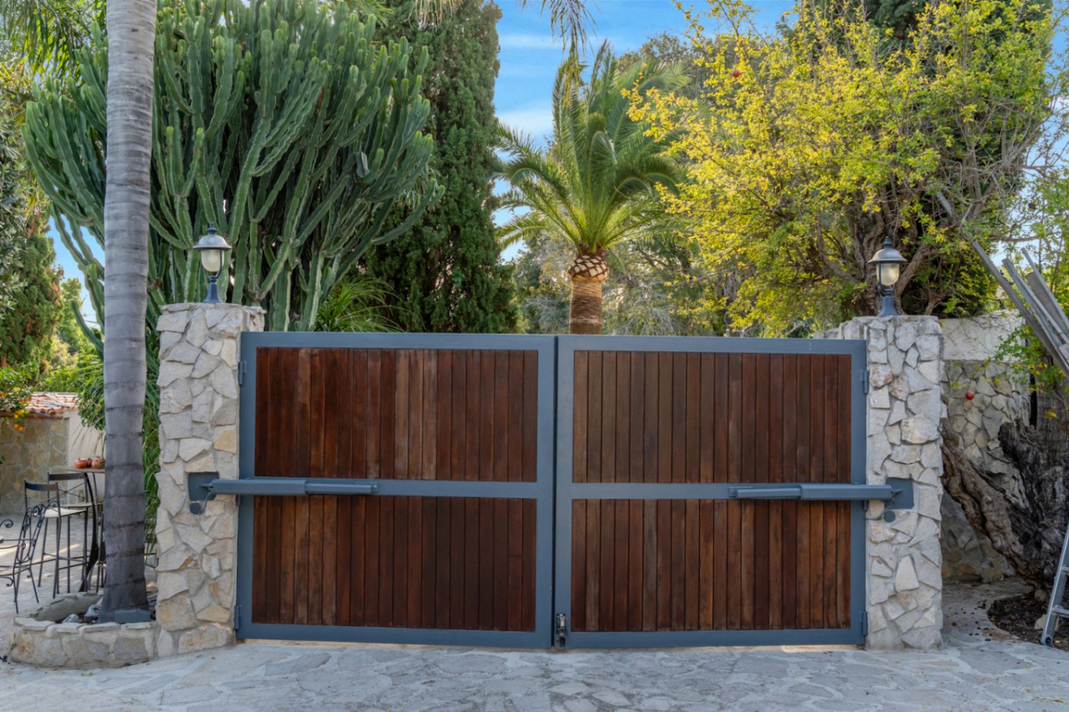 Automatisches Einfahrtstor mit Holzpaneelen und Metallrahmen, eingefasst von Natursteinpfeilern und umgeben von üppiger mediterraner Bepflanzung.