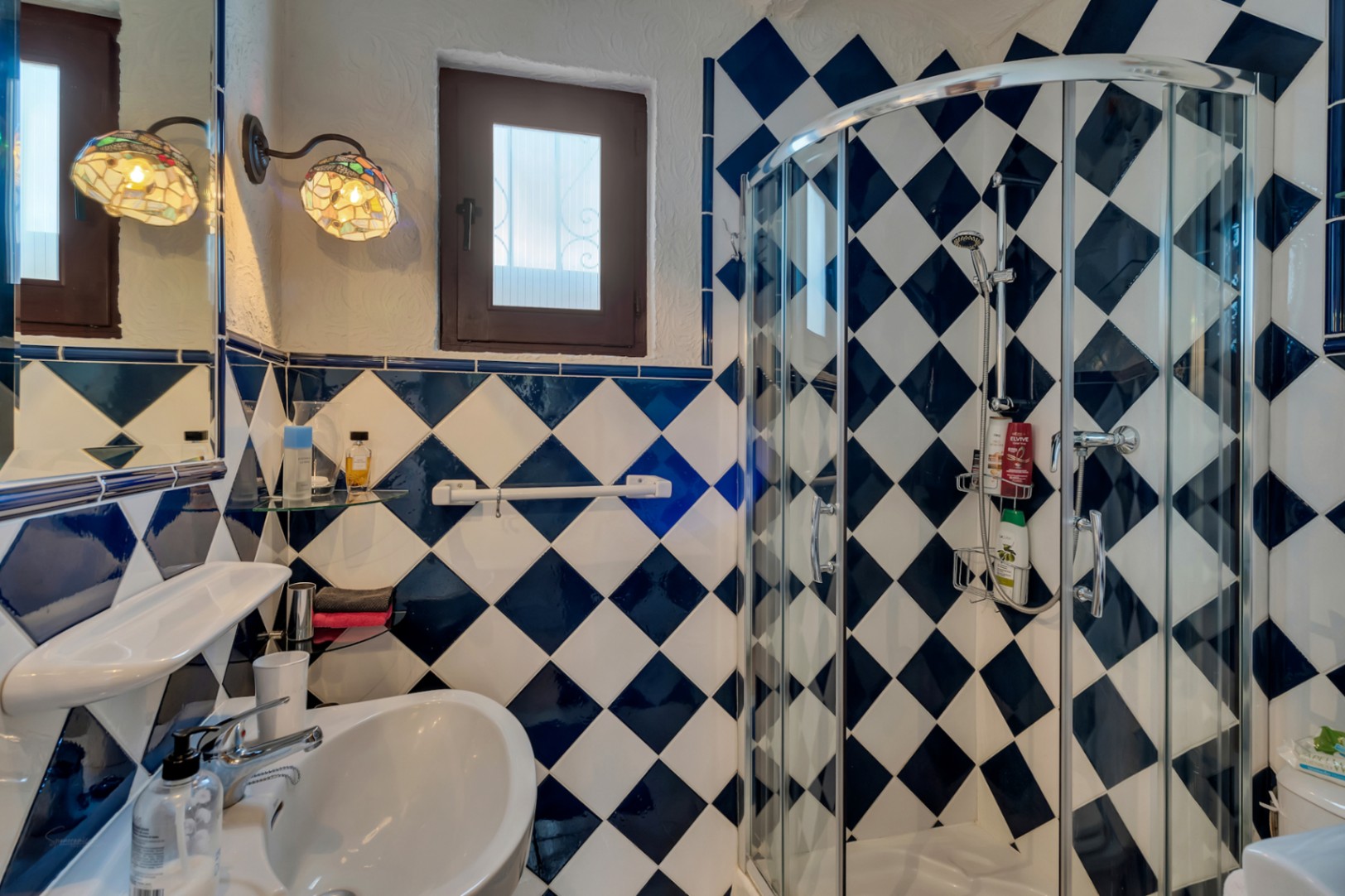 Badezimmer mit blau-weiß karierten Wandfliesen, einer gebogenen Glasduschkabine und dekorativen Wandlampen im Tiffany-Stil neben einem kleinen Holzfenster.