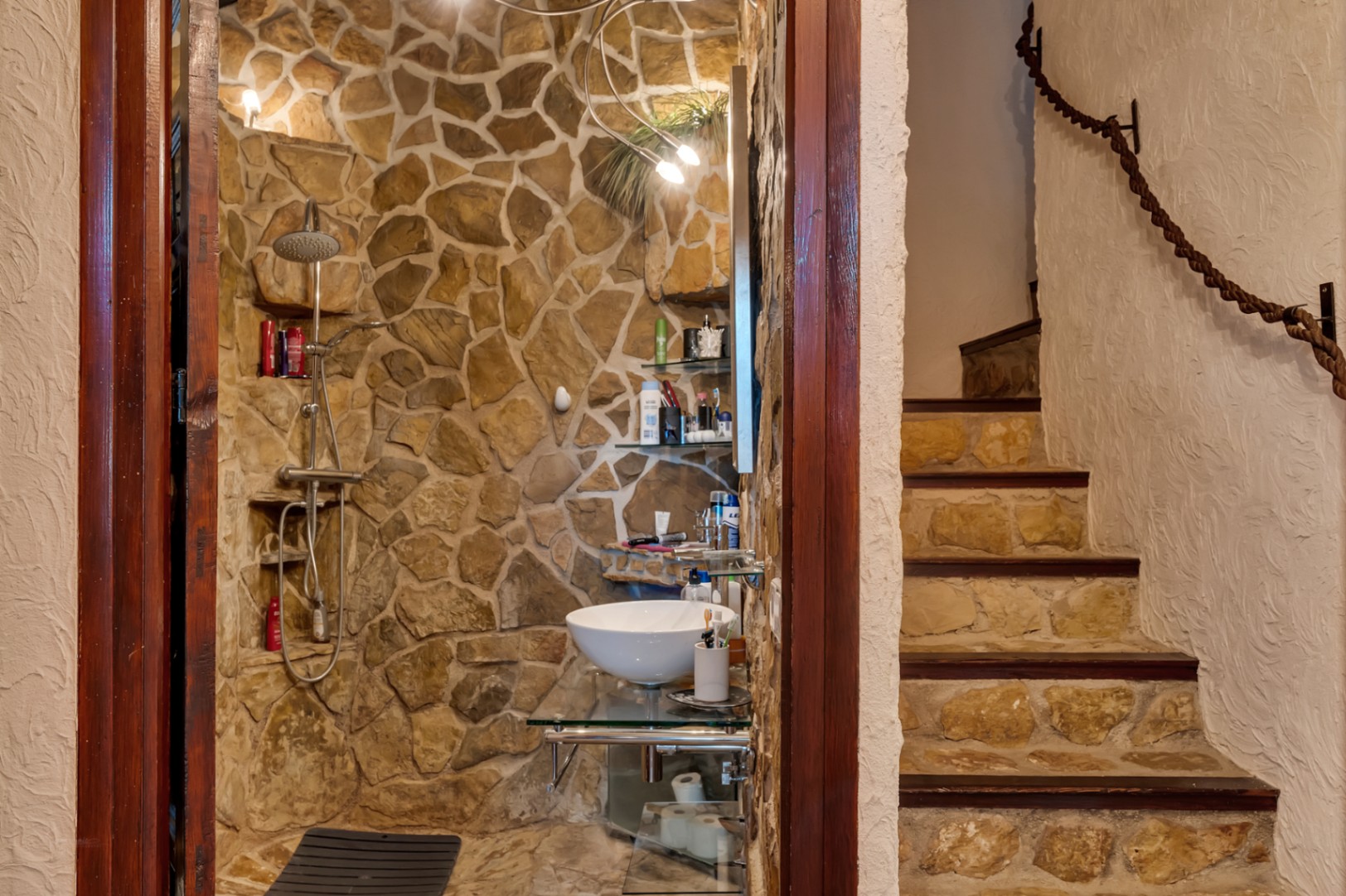 Badezimmer mit Natursteinwänden, Glaswaschtisch und integrierter Dusche. Angrenzende Treppe mit Steinstufen und einem rustikalen Seilhandlauf.