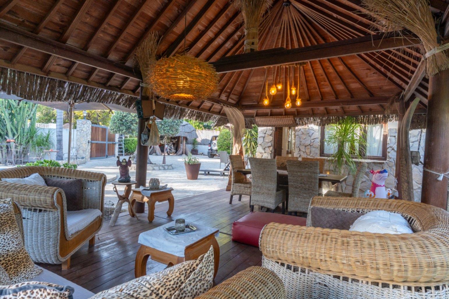 Balinesische Holzpergola mit offenem Dachstuhl, gemütlicher Lounge-Ecke aus Korbgeflecht und Essbereich auf einem dunklen Echtholzboden.