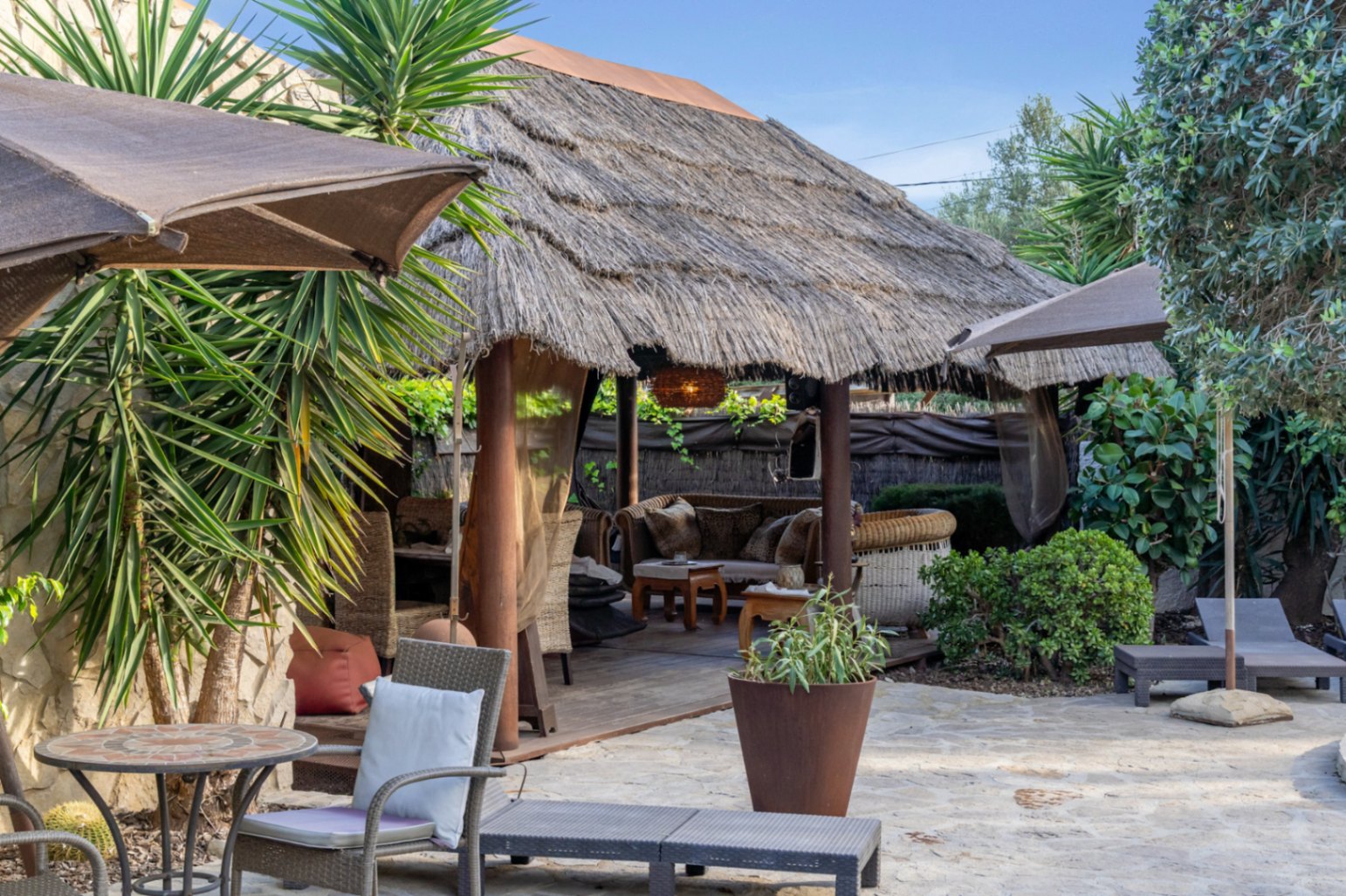 Balinesische Pergola mit Reetdach und Holzpfeilern auf einer Natursteinterrasse, umgeben von mediterraner Bepflanzung und komfortablen Gartenmöbeln.