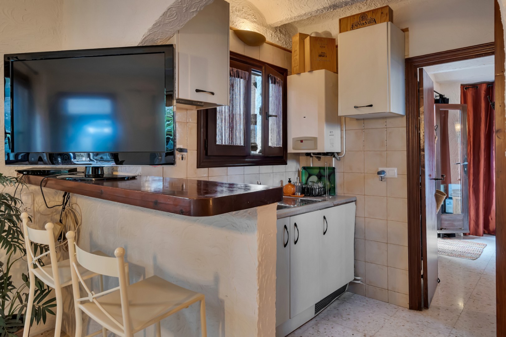 Kompakte Küchenzeile mit Holz-Frühstücksbar, weißen Schränken und integriertem Fernseher. Die Wände sind in rustikalem Strukturputz gehalten.