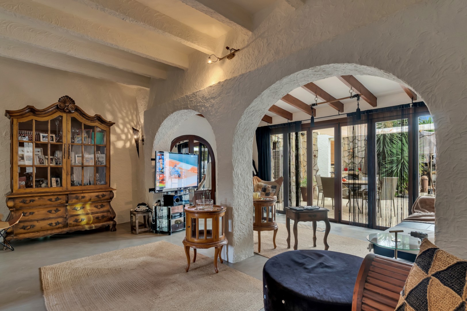 Mediterraner Wohnbereich mit strukturierten Rundbögen, Holzbalkendecke und einer klassischen Vitrine. Große Glastüren führen direkt zum angrenzenden Außenbereich.