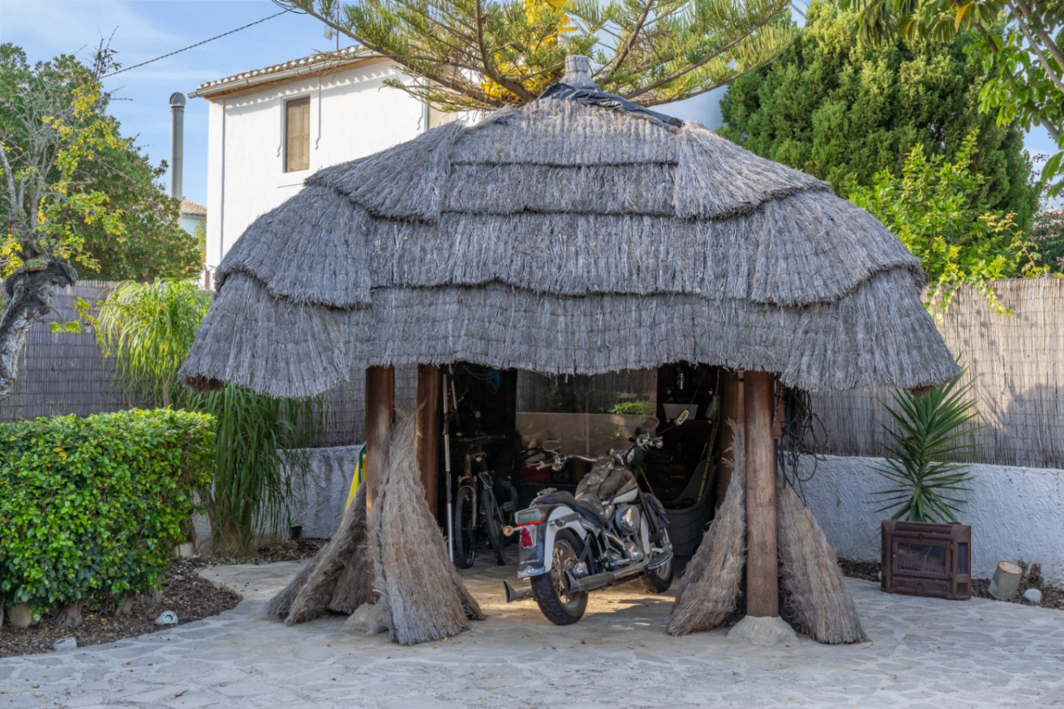 Pergola im balinesischen Stil mit dickem Reetdach und Holzpfosten, die als überdachter Stellplatz für Motorräder und Fahrräder auf Natursteinpflaster dient.